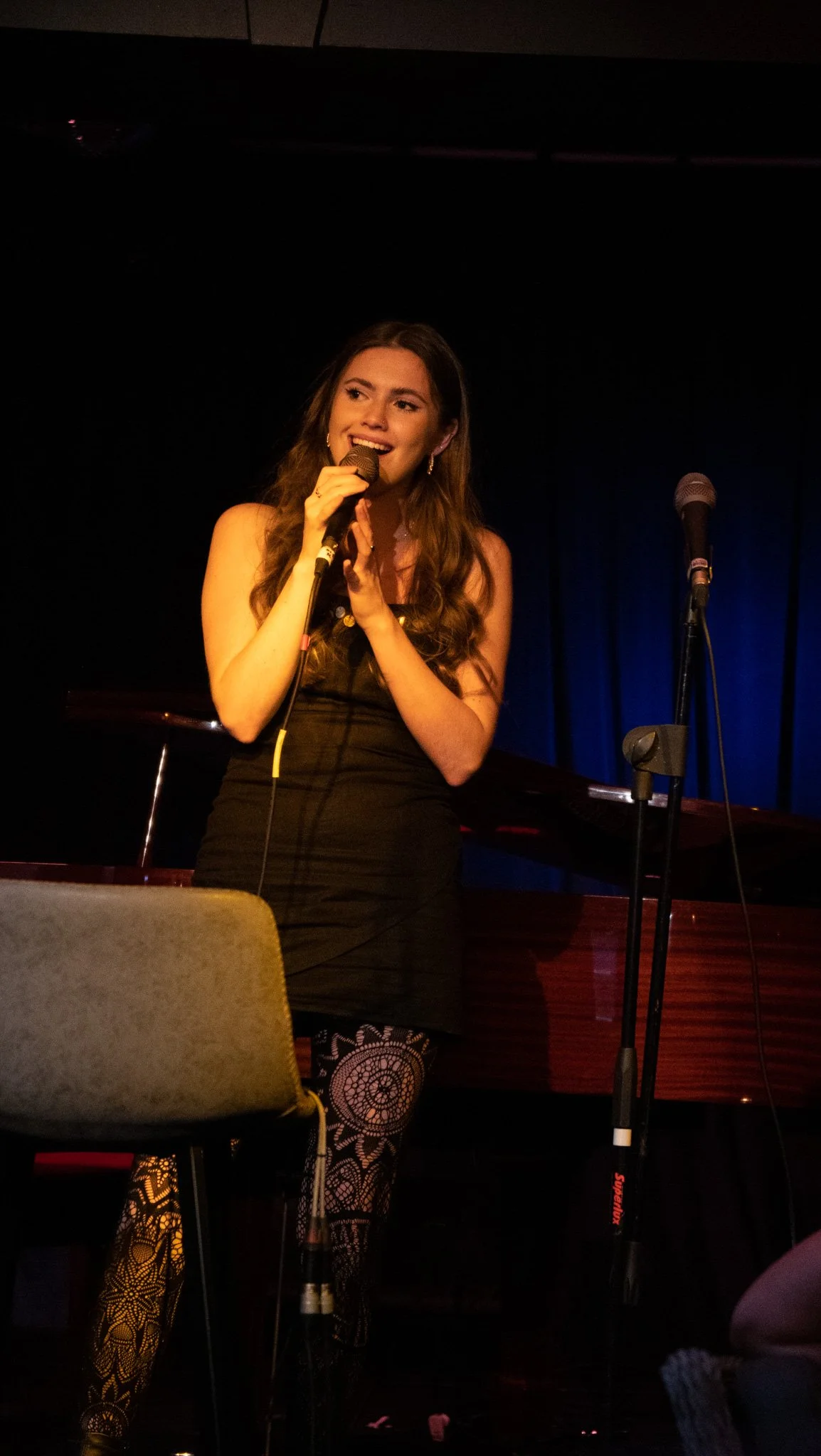 Young woman singing into a microphone on stage with a piano and microphone stand behind her. She has long brown hair and is wearing a black dress, patterned tights, and earrings, in a dimly lit performance space.