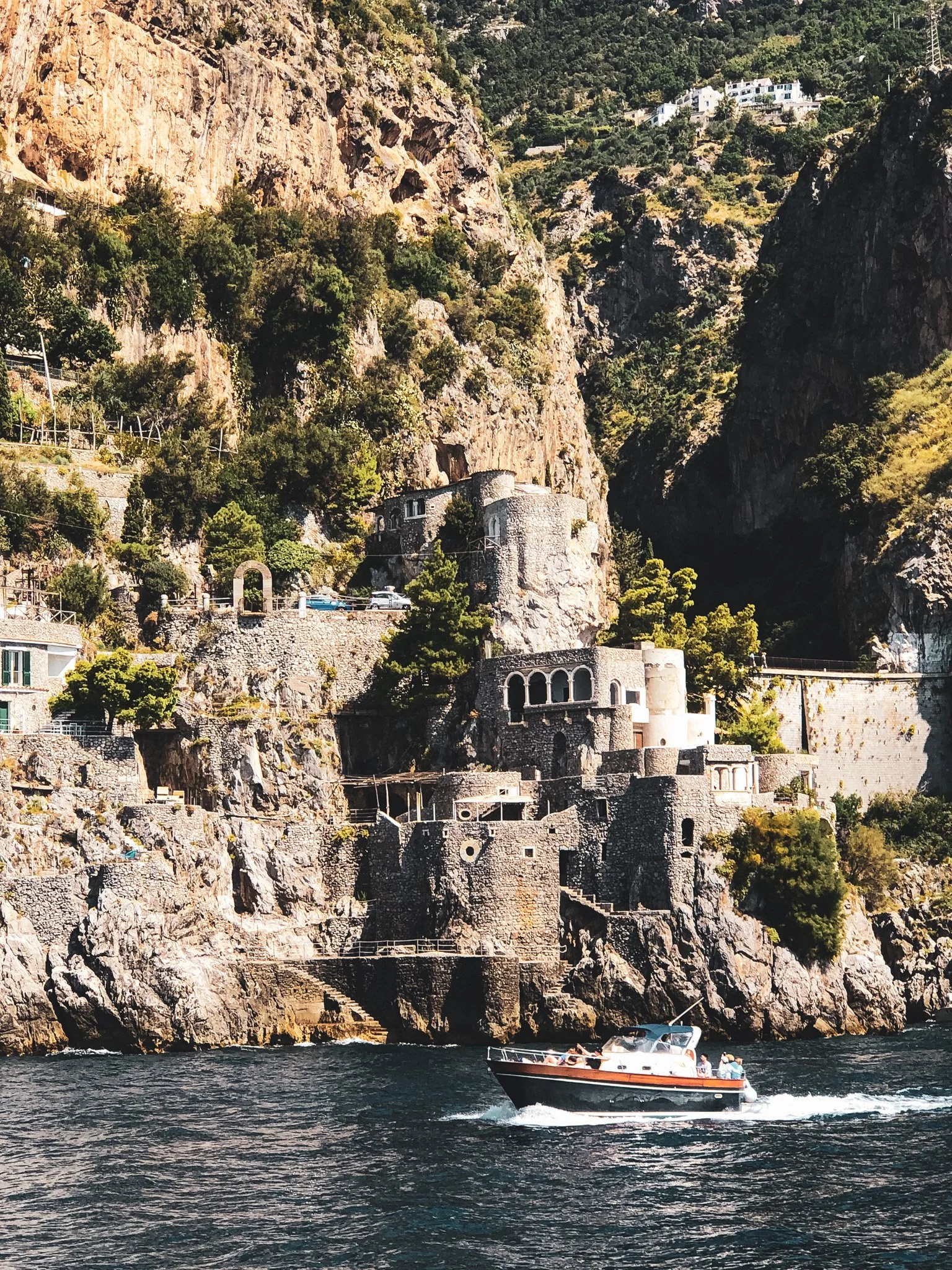 A boat with passengers sailing near the rocky coast of a hillside with houses and greenery, under a sunny sky.