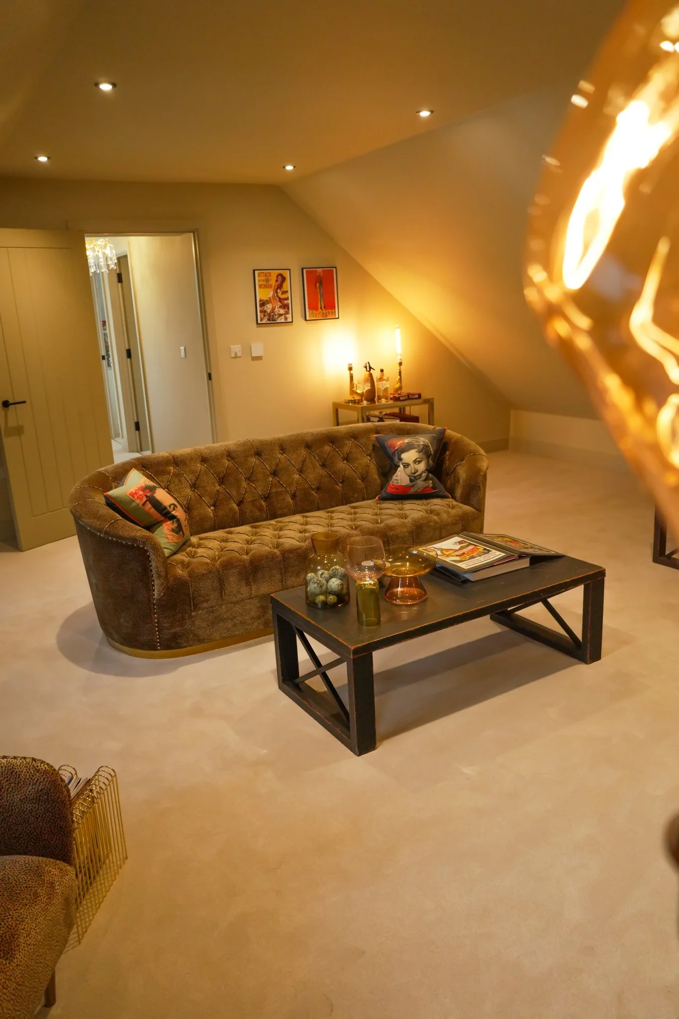 Living room with beige carpet, a tufted brown velvet sofa with pillows featuring women on pins, a black coffee table with decorative vases and magazines, beige walls with framed posters, a small table with bottles and candles, slips to other rooms, and ceiling lights.