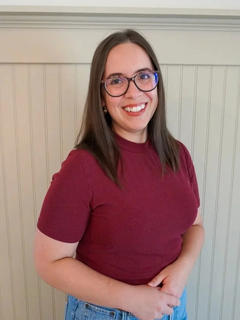 A woman with long brown hair, glasses, and hoop earrings smiling while wearing a maroon shirt and blue jeans, standing in front of a beige wainscoted wall.