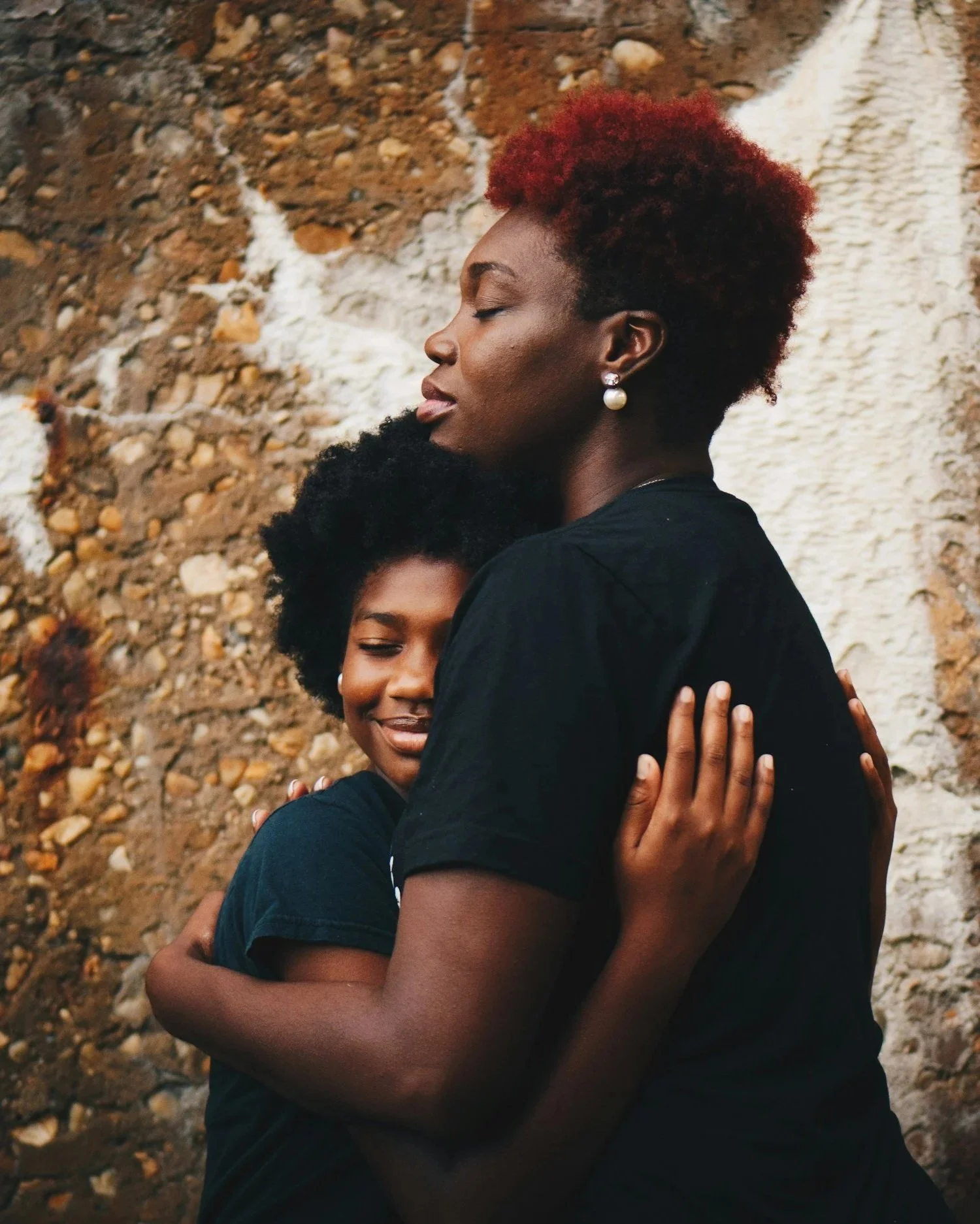Two women hugging each other with closed eyes against a rocky background, one with short curly black hair and the other with short red hair.