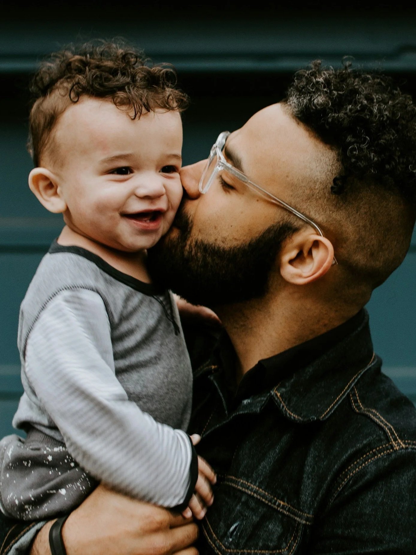 A man with glasses and a beard holding a smiling young child with curly hair, close to his face.