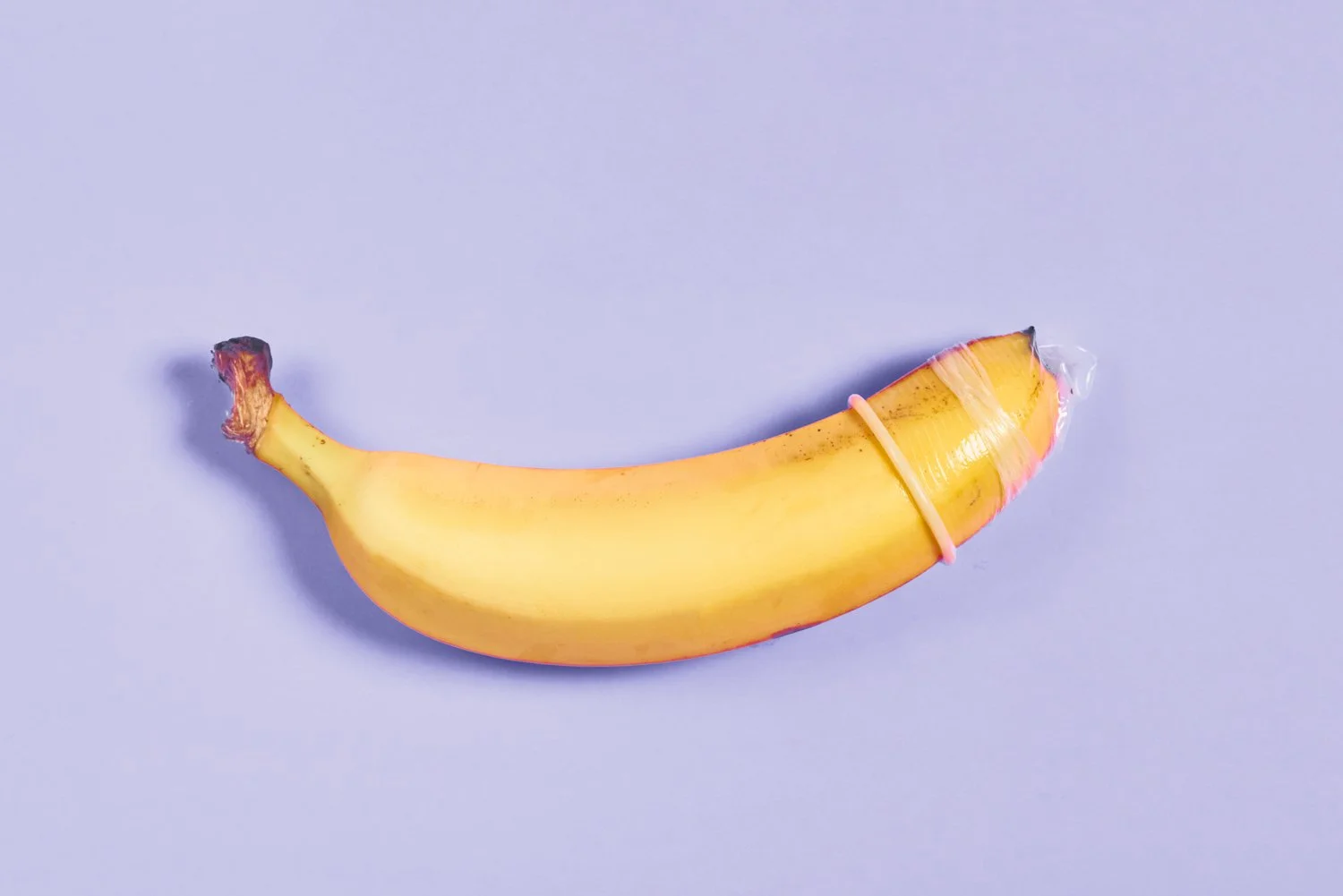 A banana with a small rubber band around its middle on a light purple background.