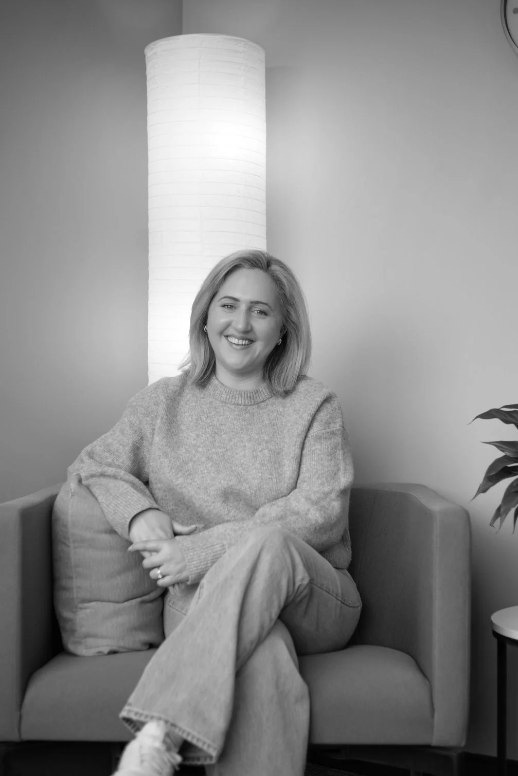 A woman with shoulder-length hair sitting on a sofa, smiling at the camera, in a room with a tall floor lamp and a plant visible on the right.