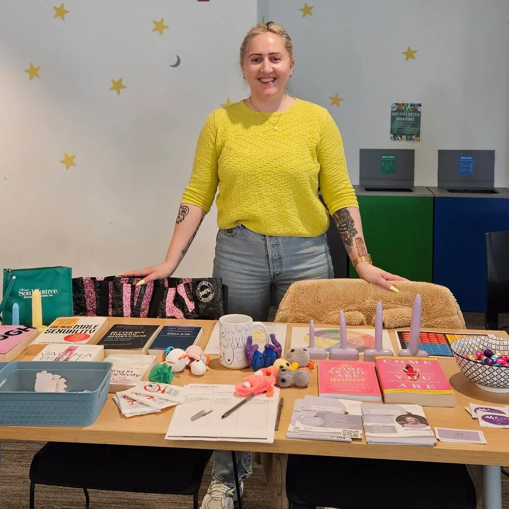 A smiling woman standing behind a table filled with various items, including books, plush toys, candles, and pamphlets, in a room decorated with star and moon wall decals.