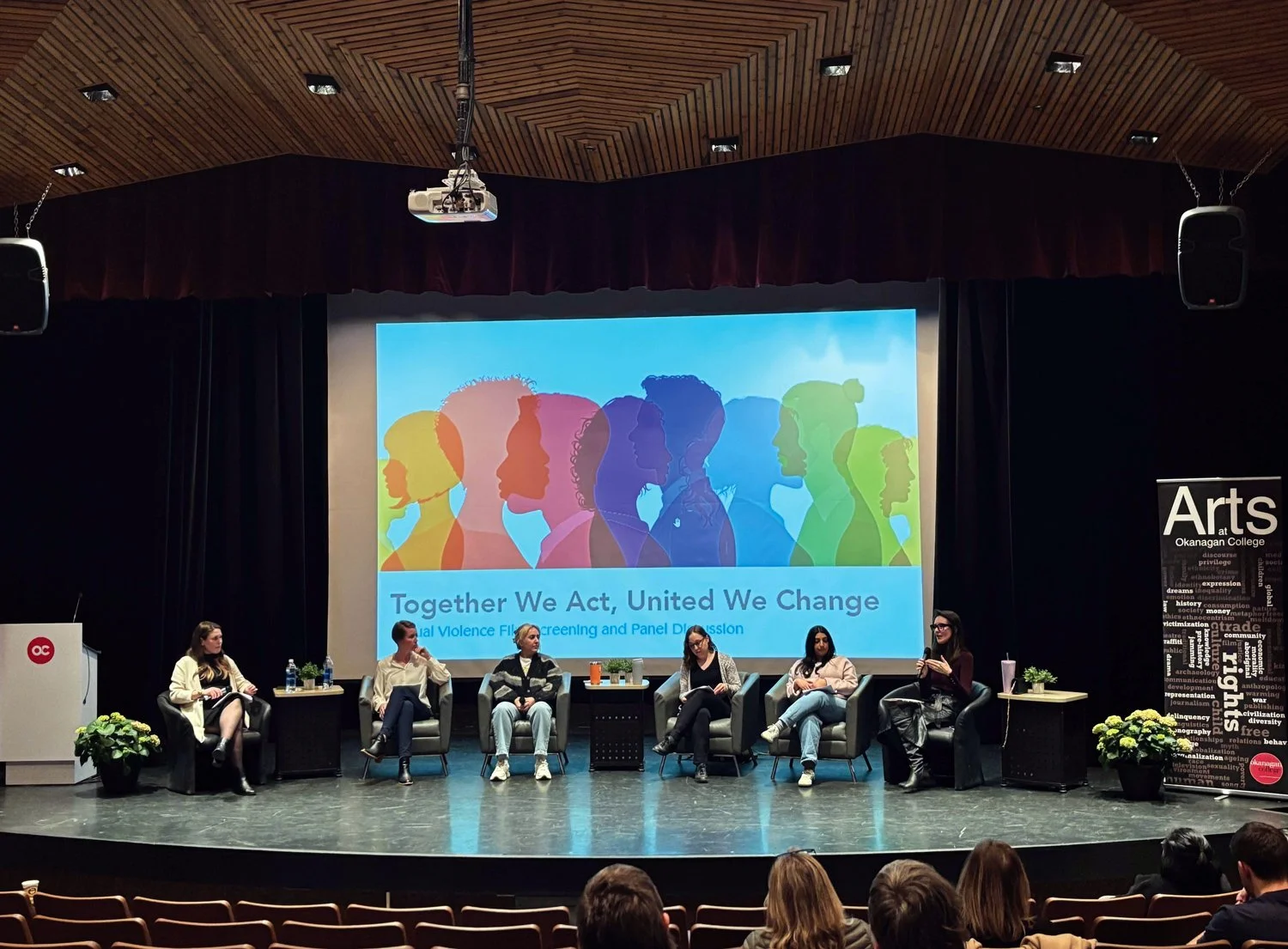 Panel discussion on stage at Okanagan College with a large screen displaying rainbow-colored silhouettes of diverse women and the text 'Together We Act, United We Change.' Six women sit in chairs on stage, speaking and listening, with potted plants on side tables, and an audience watching.