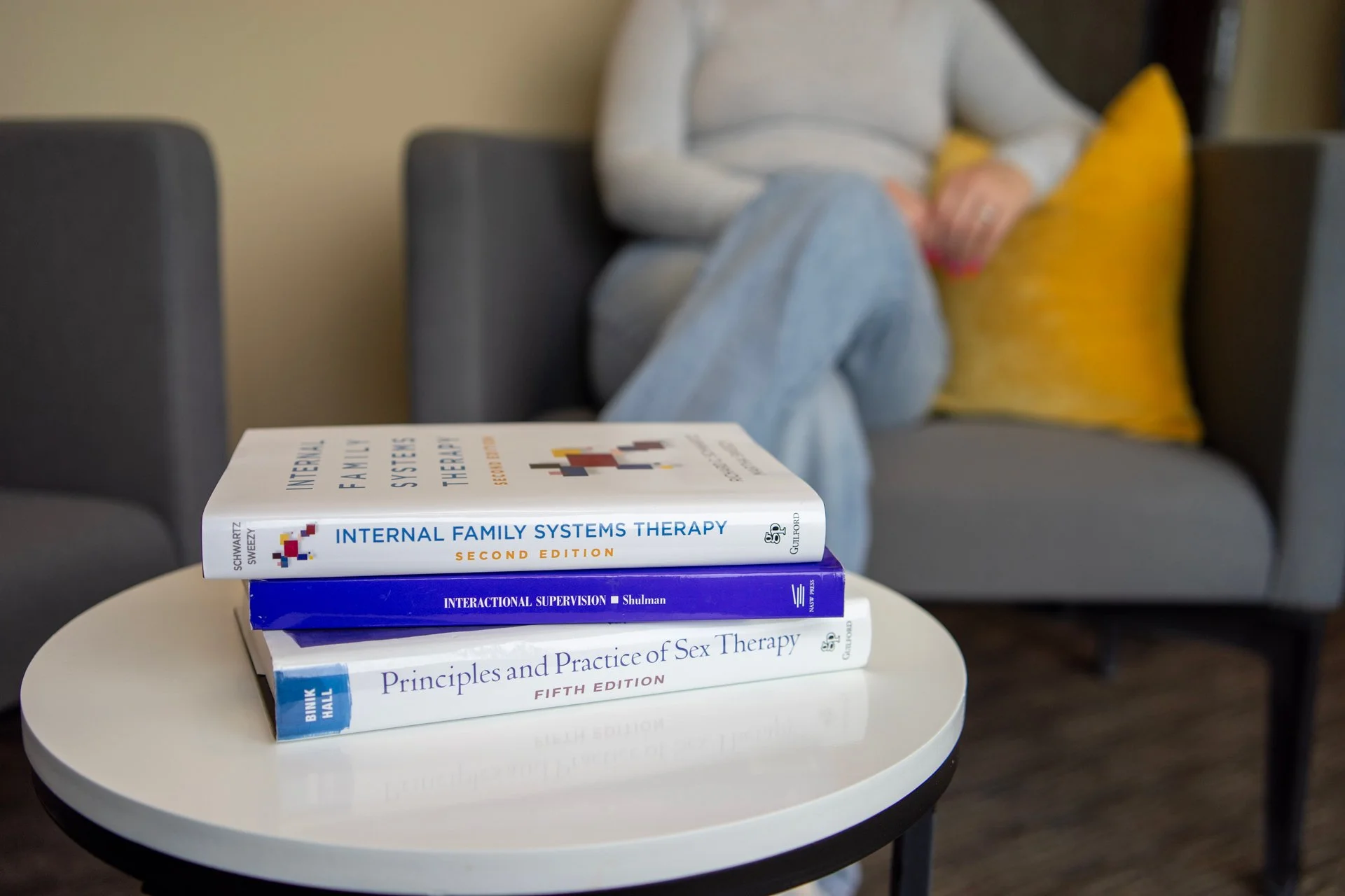 Stack of four psychology and therapy books on a round white table in front of a person sitting on a gray couch with a yellow pillow, blurred background.