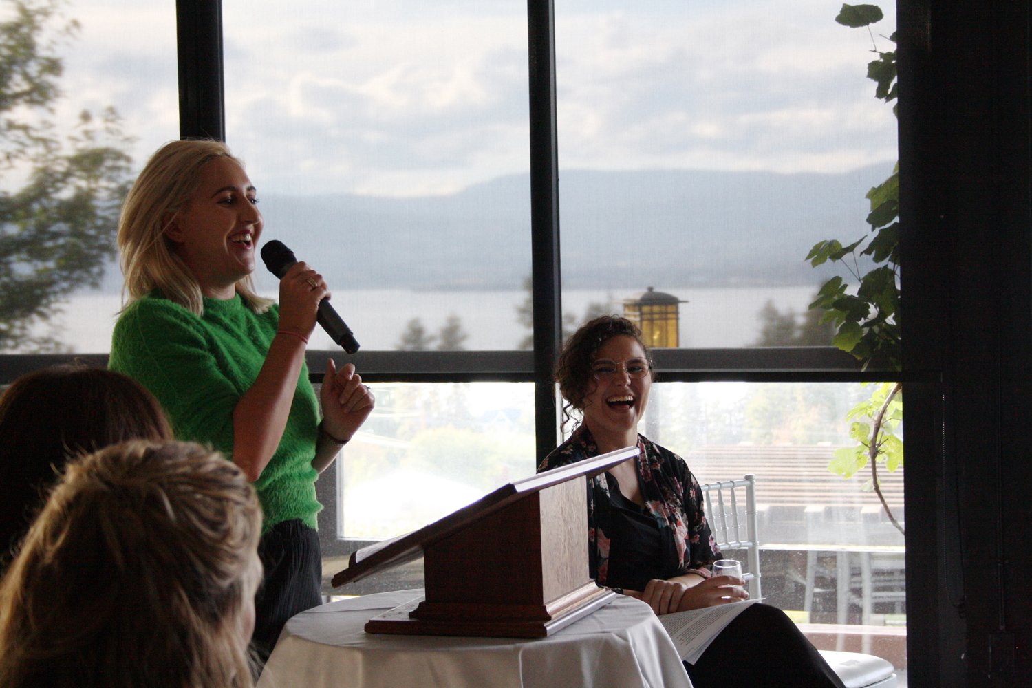 A woman in a green sweater is speaking into a microphone during an event. Another woman is seated next to her, laughing and holding a glass, with an open book on her lap. The event is taking place indoors with a large window showing an outdoor view of trees, water, and mountains.