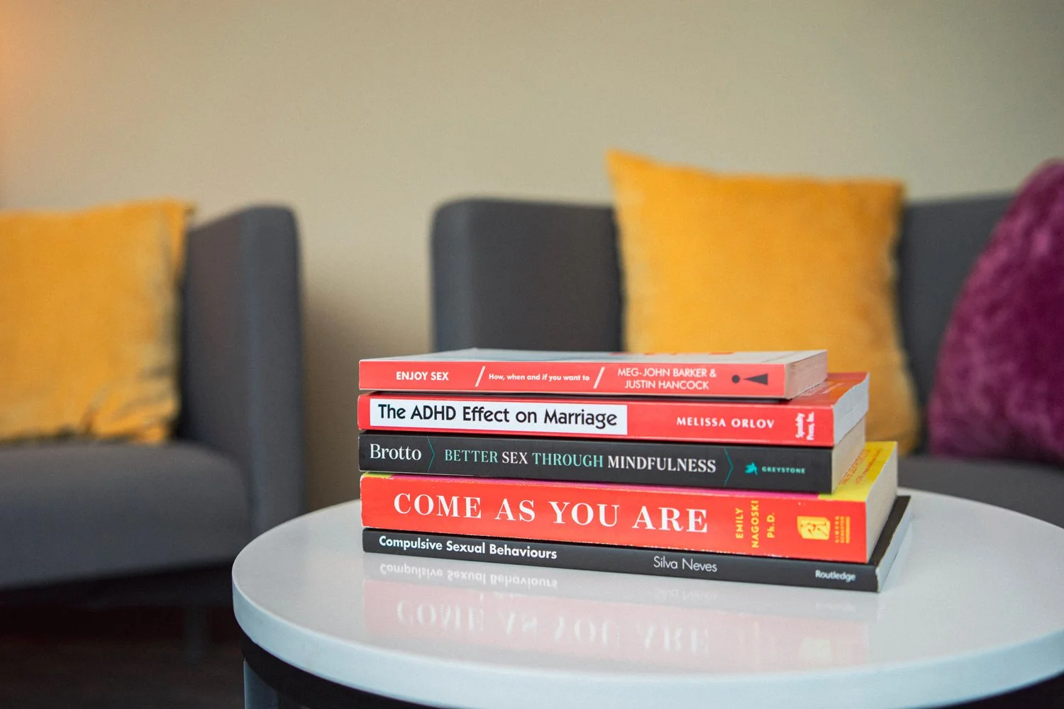 A stack of five books on a white round table with gray chairs and yellow pillows in the background.