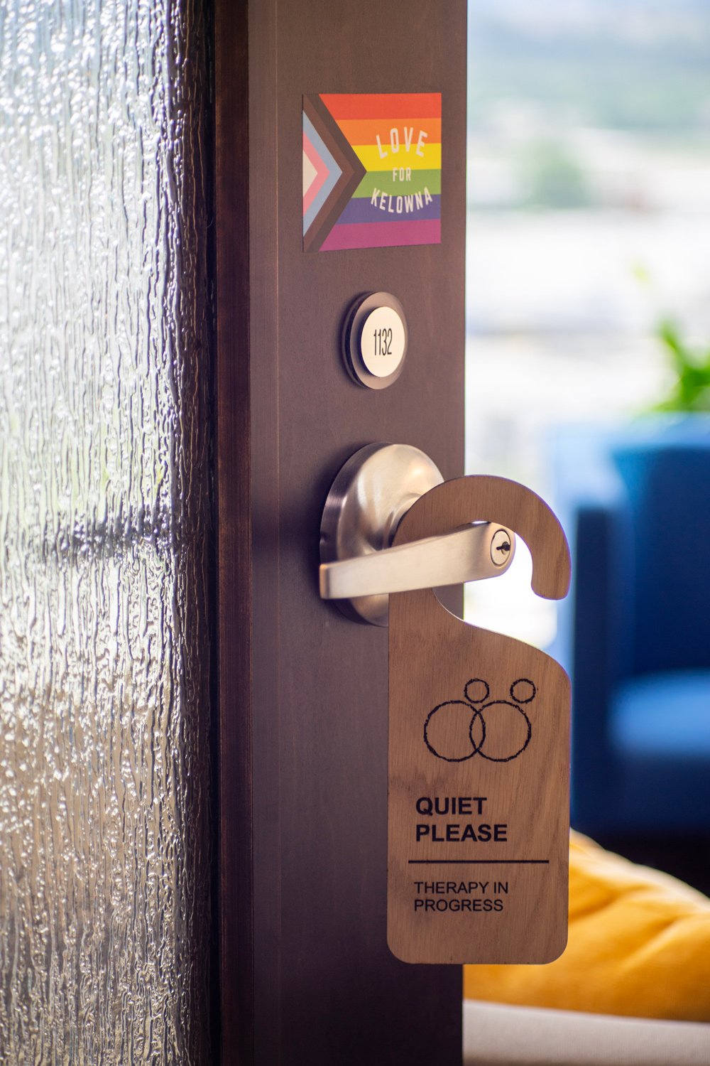 A door with a wood-colored "Quiet Please" sign indicating therapy in progress. There is a door handle, a small round button with the number 1132, and a sticker with rainbow colors and the words "Love for Kelowna."