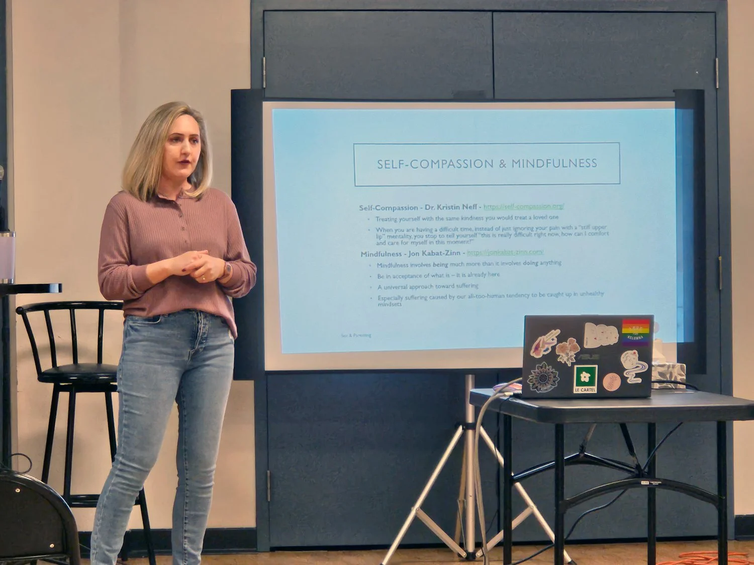 Woman giving a presentation on self-compassion and mindfulness in a room with a large screen displaying her slides.