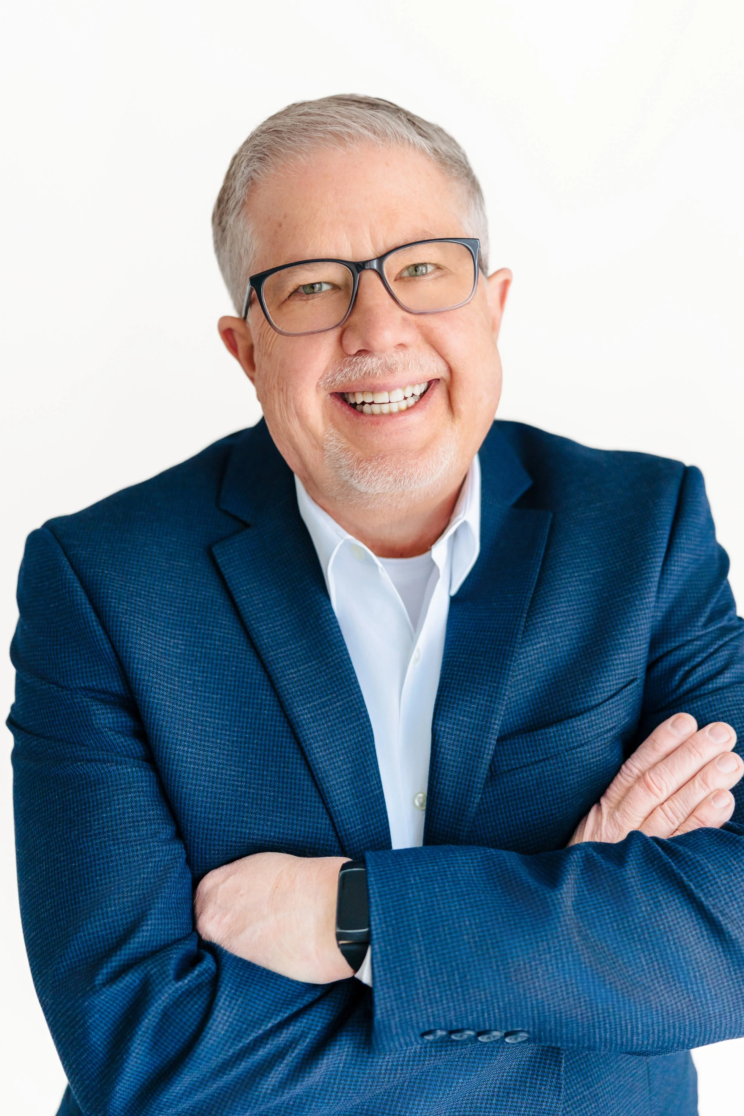 A middle-aged man with gray hair, glasses, and a goatee, smiling and wearing a navy blue suit jacket with a white shirt, standing against a white background.