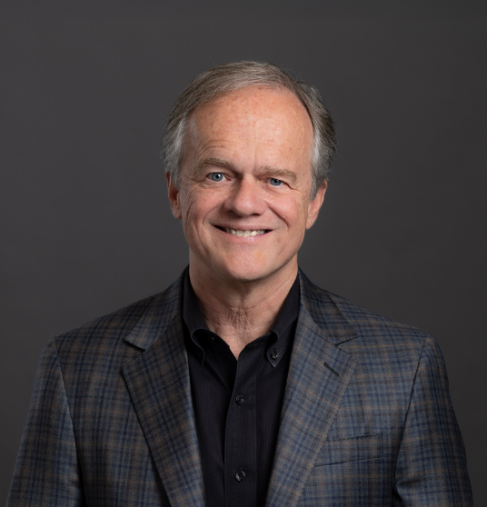 Portrait of a middle-aged man with light skin, gray hair, and a friendly smile, wearing a dark plaid blazer and a black shirt, standing against a plain dark gray background.