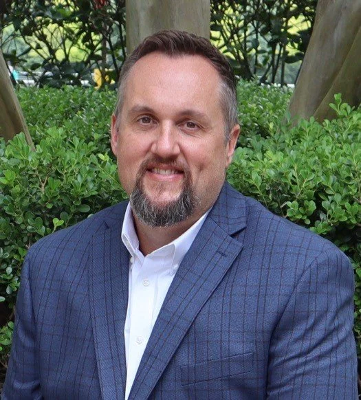 A man with a beard and mustache wearing a blue checkered suit and white shirt, smiling outdoors in front of green bushes and trees.