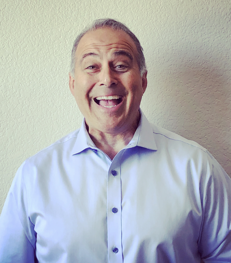 A smiling middle-aged man with short gray hair, wearing a light blue dress shirt, standing against a neutral wall background.