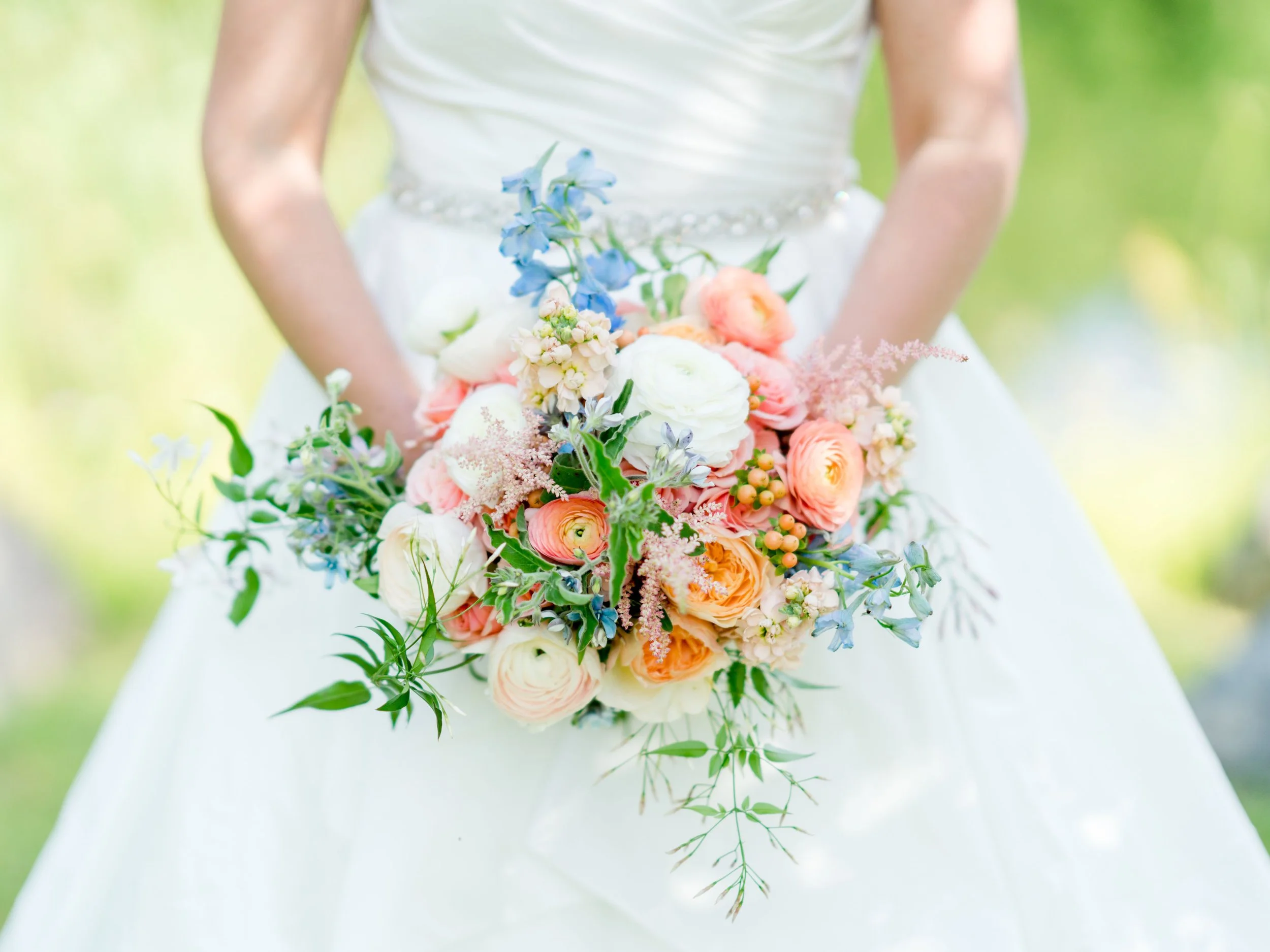 A bride holding a colorful bouquet of flowers with peach, white, and blue blossoms, against a blurred green outdoor background.