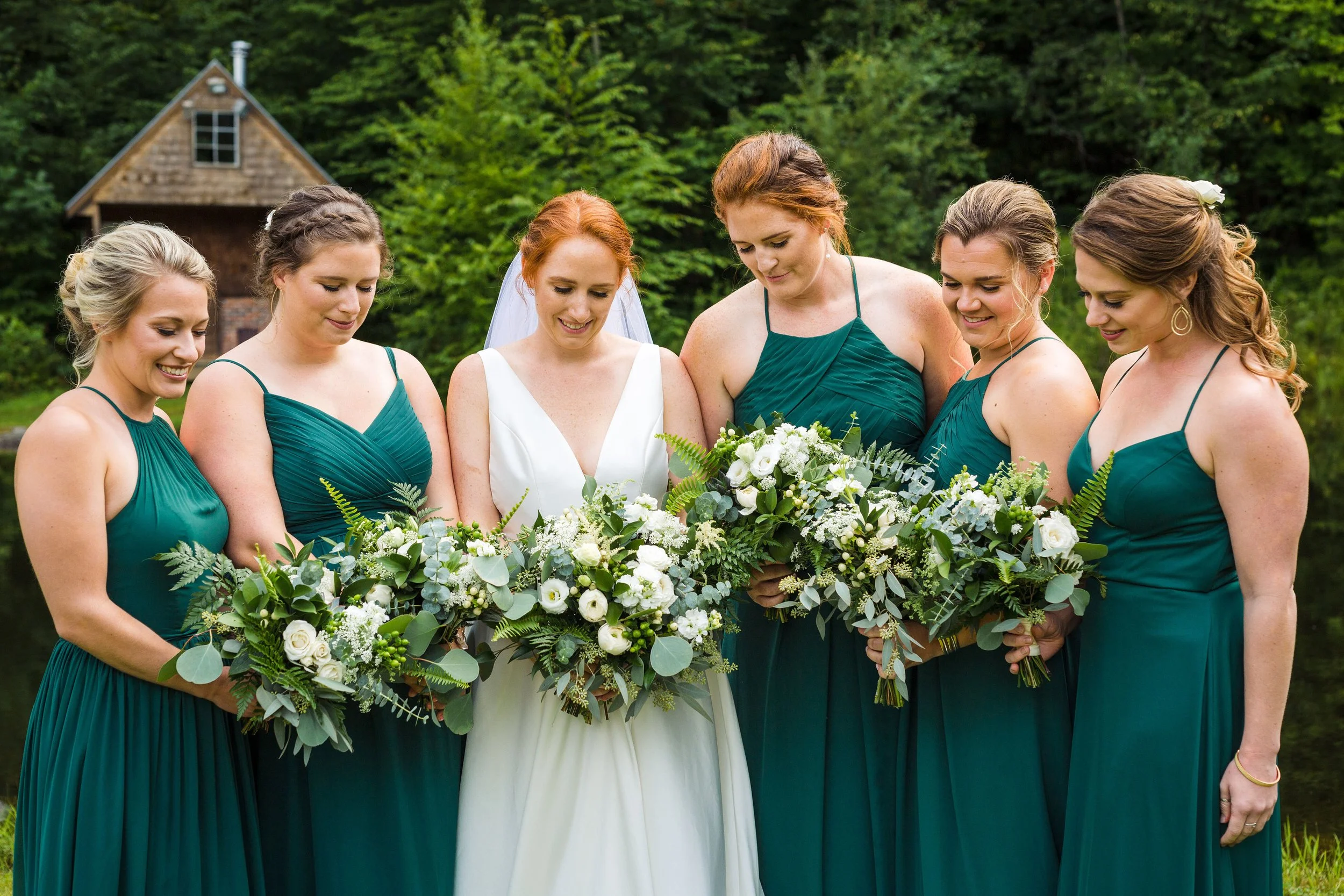 A bride and her bridesmaids stand together outdoors, holding a large bouquet of white flowers and greenery, with a rustic house and trees in the background.