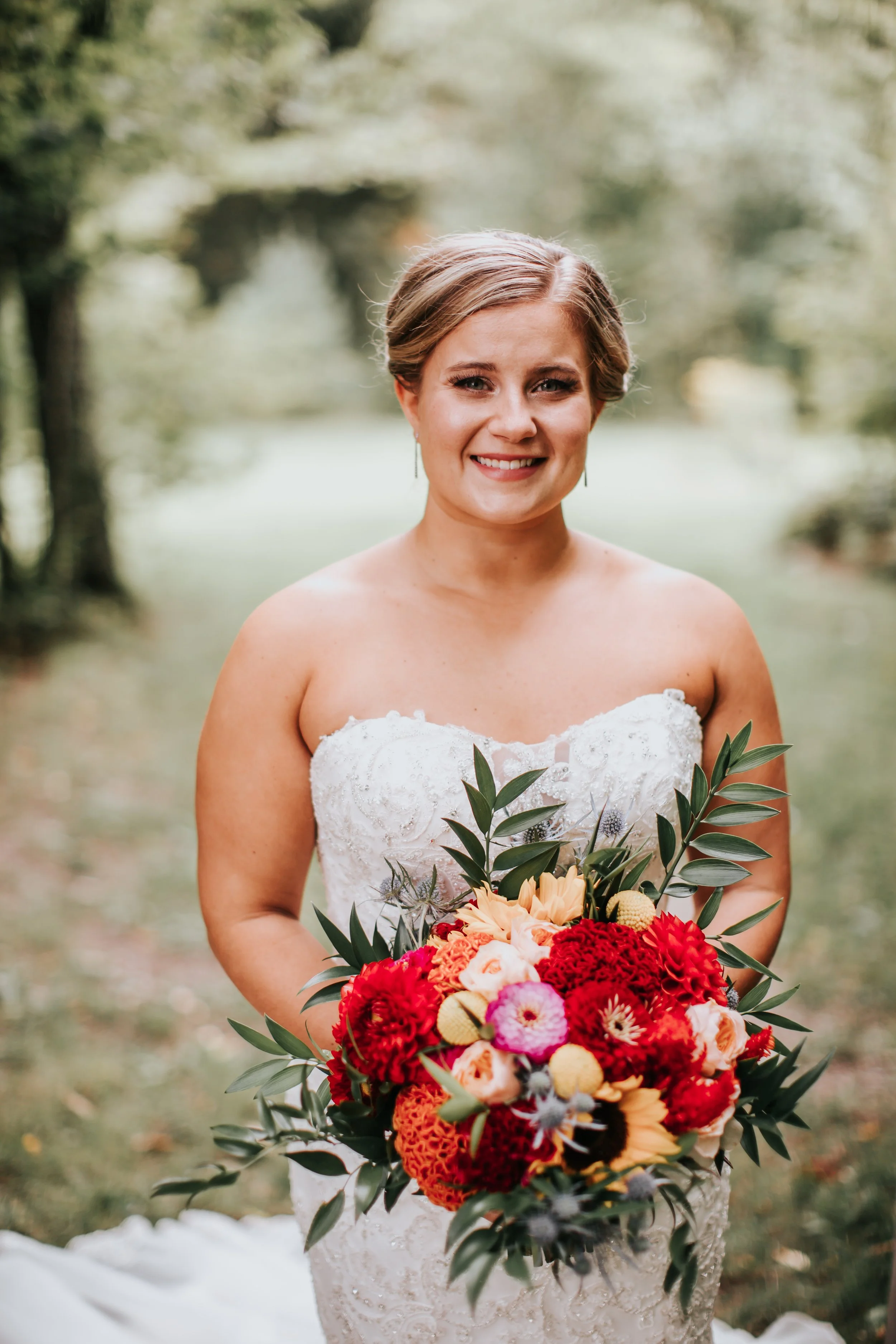 A smiling bride in a strapless white wedding dress holding a colorful bouquet of red, pink, orange, and yellow flowers with green leaves, outdoors with trees in the background.