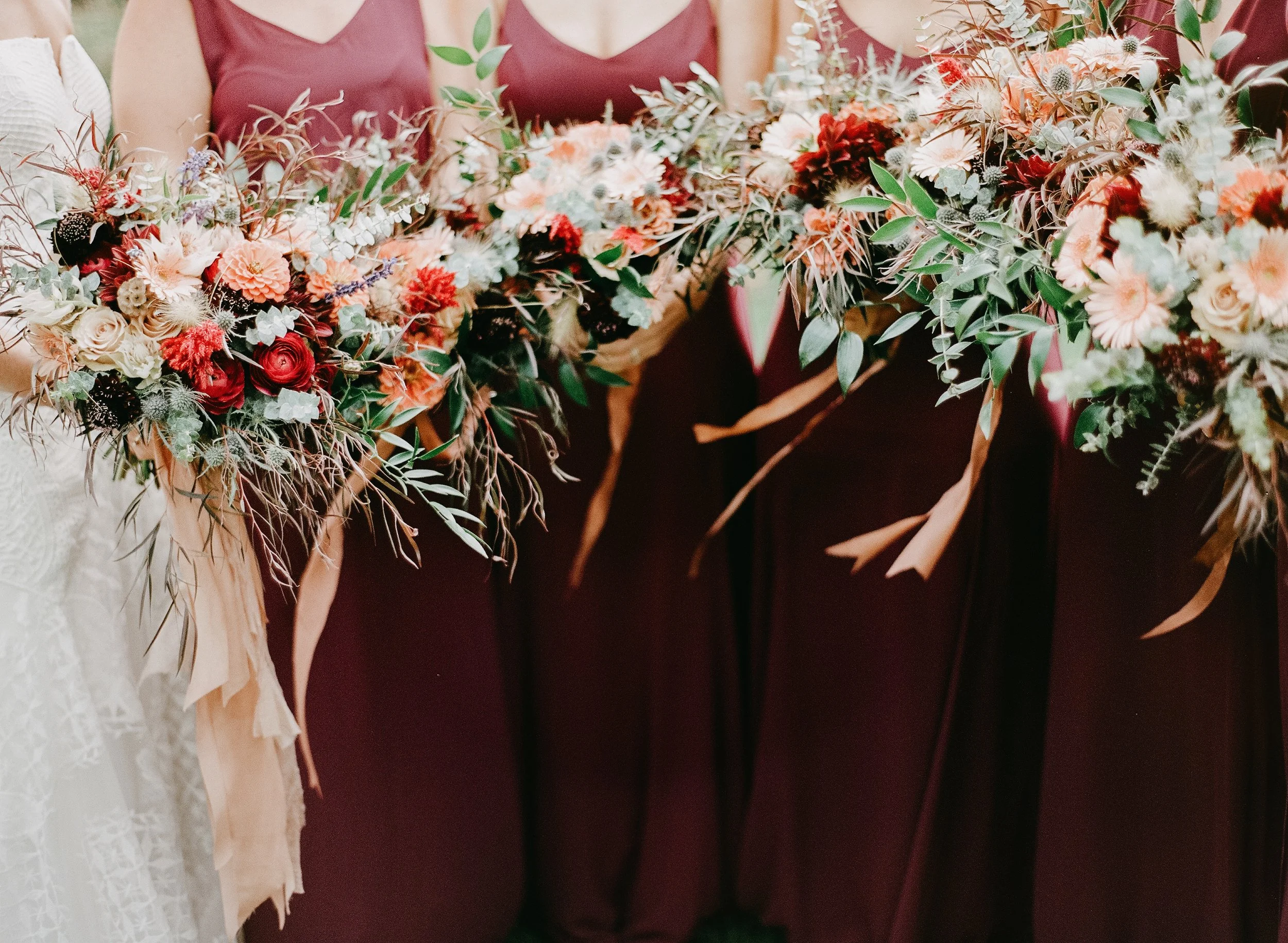 Close-up of a floral arrangement with peach, red, and white flowers, and greenery, held by people dressed in formal attire.