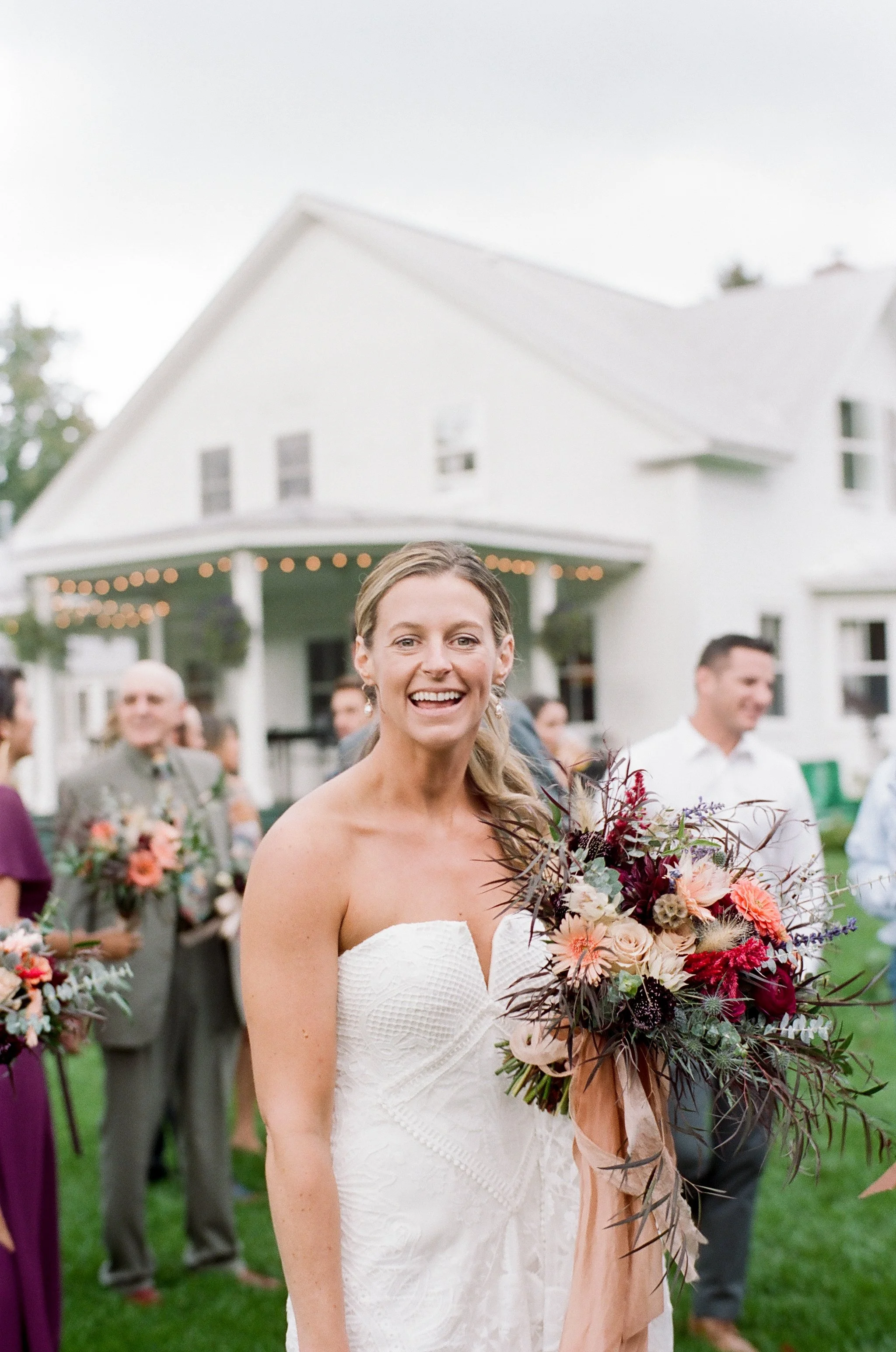 A smiling woman in a white strapless wedding dress holding a bouquet of pink, purple, and red flowers at an outdoor wedding gathering.
