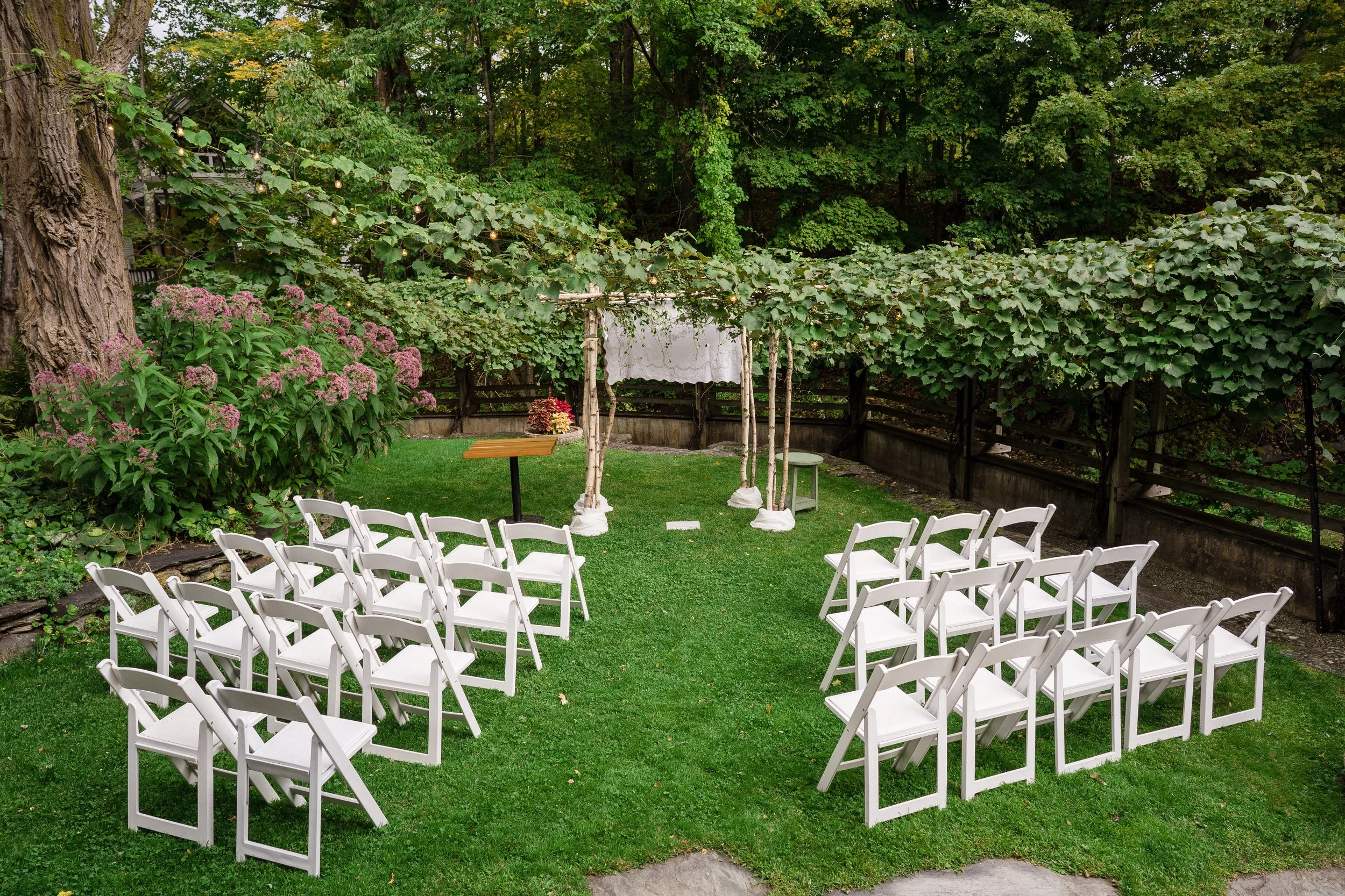 Outdoor wedding ceremony setup with white chairs arranged on a green lawn, a wooden wedding arch draped with white fabric, surrounded by lush green trees and flowering bushes.