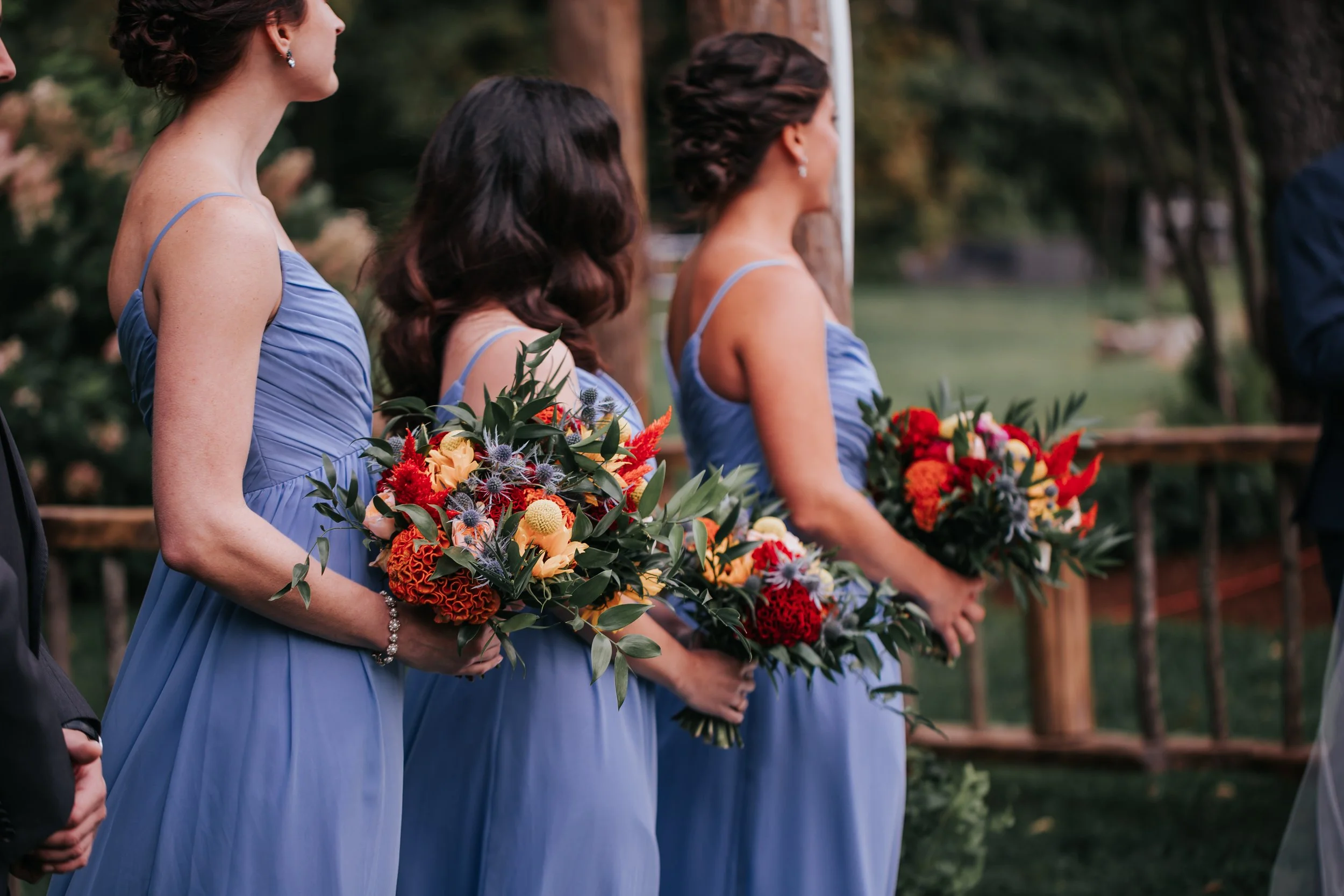 Women in blue dresses holding colorful flower bouquets at an outdoor event, possibly a wedding ceremony.