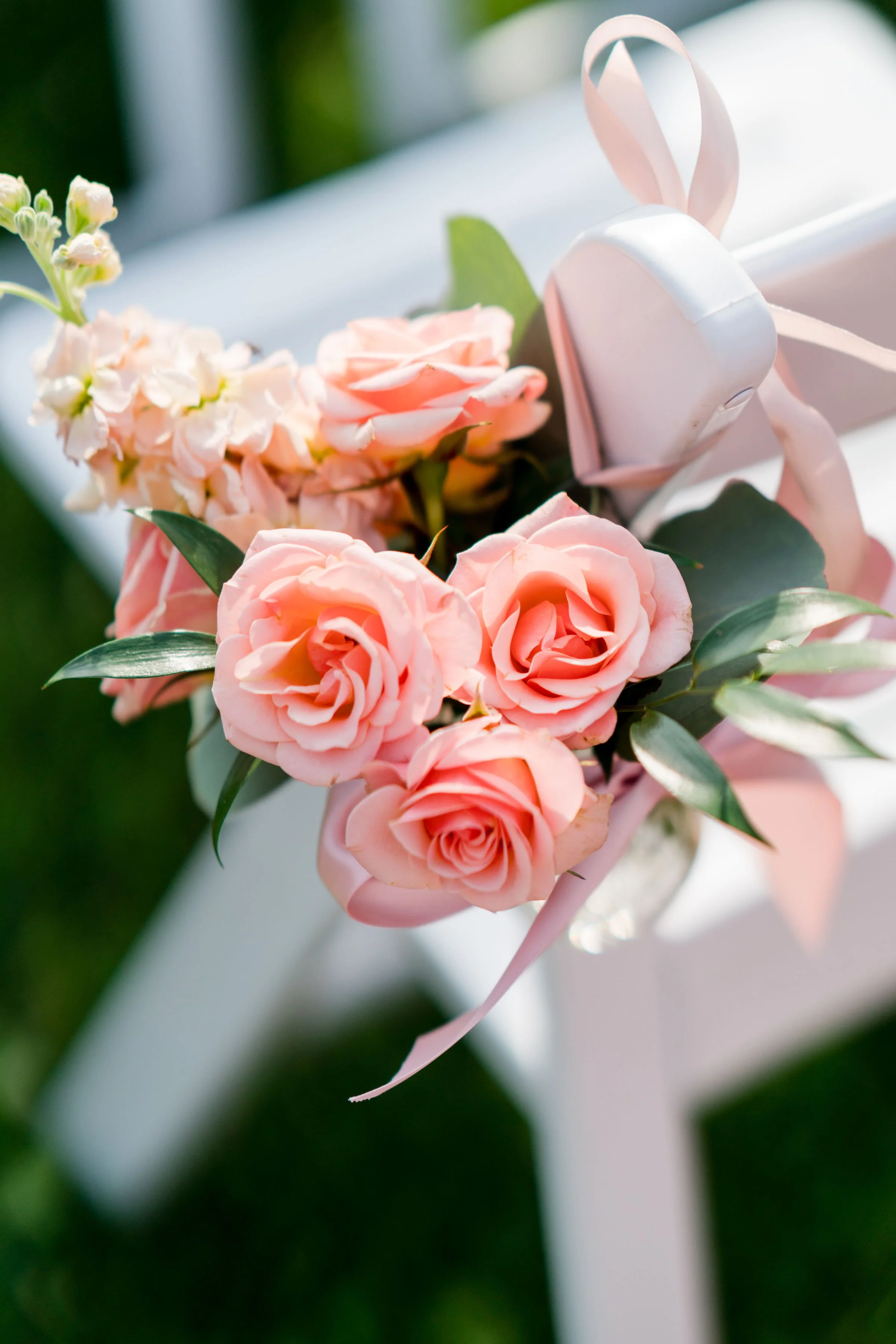 A close-up of a bouquet of pink roses and small white flowers with green leaves, tied with a pink ribbon, attached to a white pole or stake.