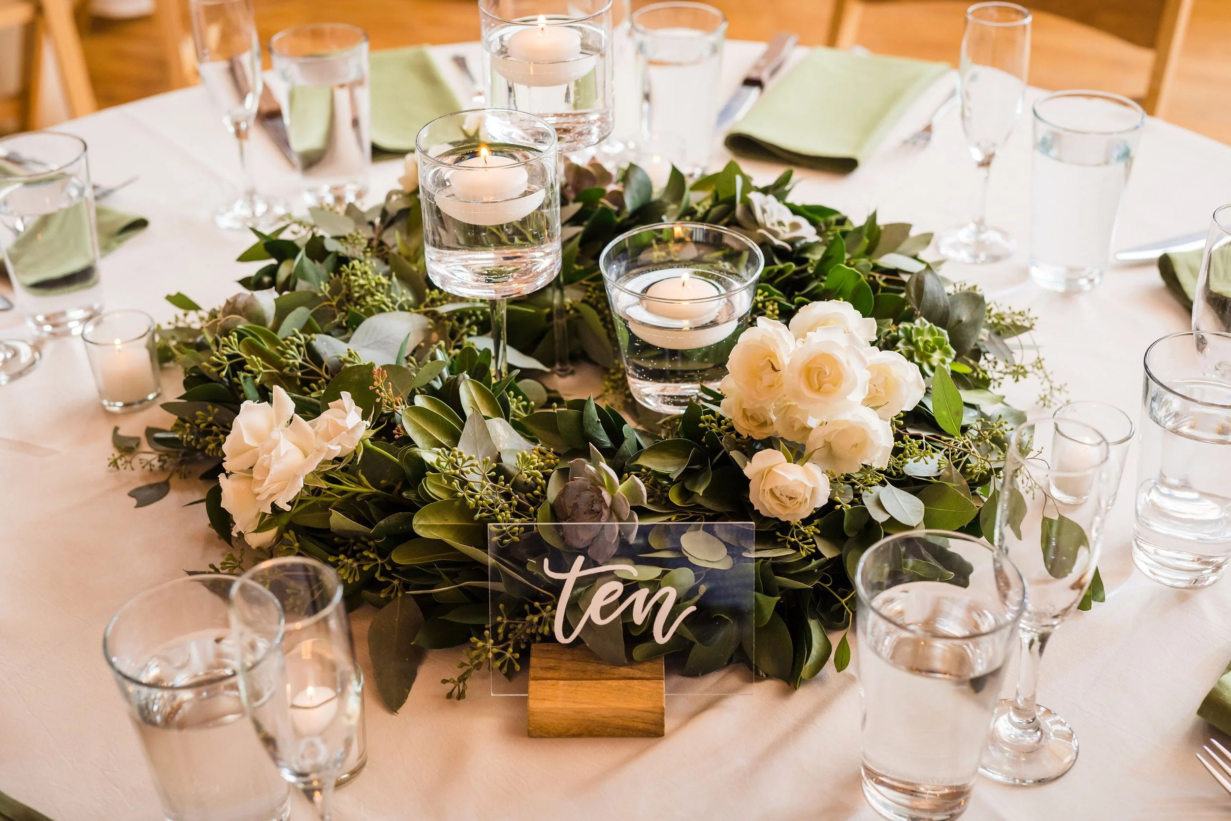 A round table decorated with a floral centerpiece of white roses and green leaves, surrounded by empty water glasses and green napkins, with lit floating candles and a sign that reads 'Team' in cursive.