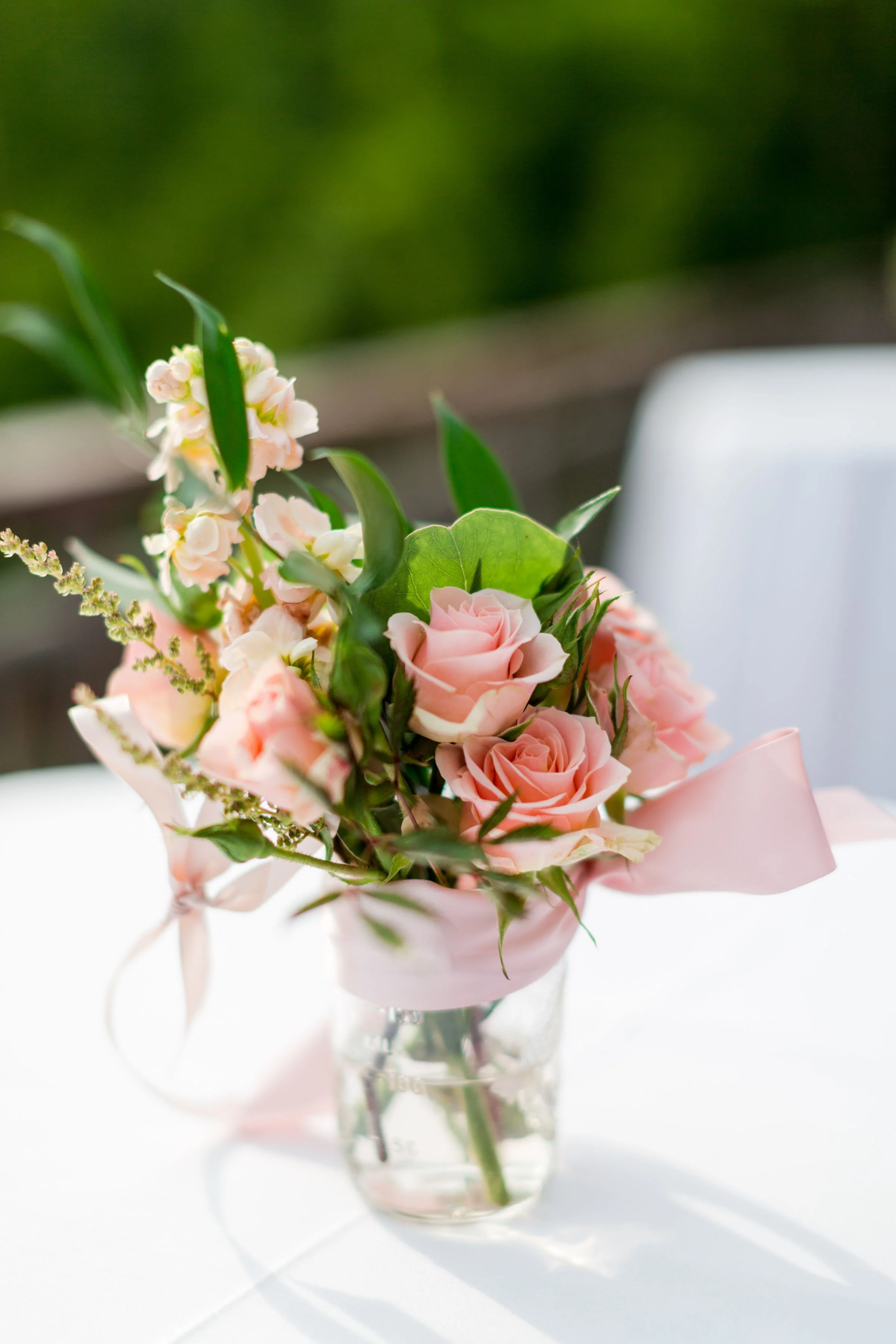 A small bouquet of pink and white roses, green leaves, and other flowers in a clear glass vase, with a pink ribbon tied around the bouquet.