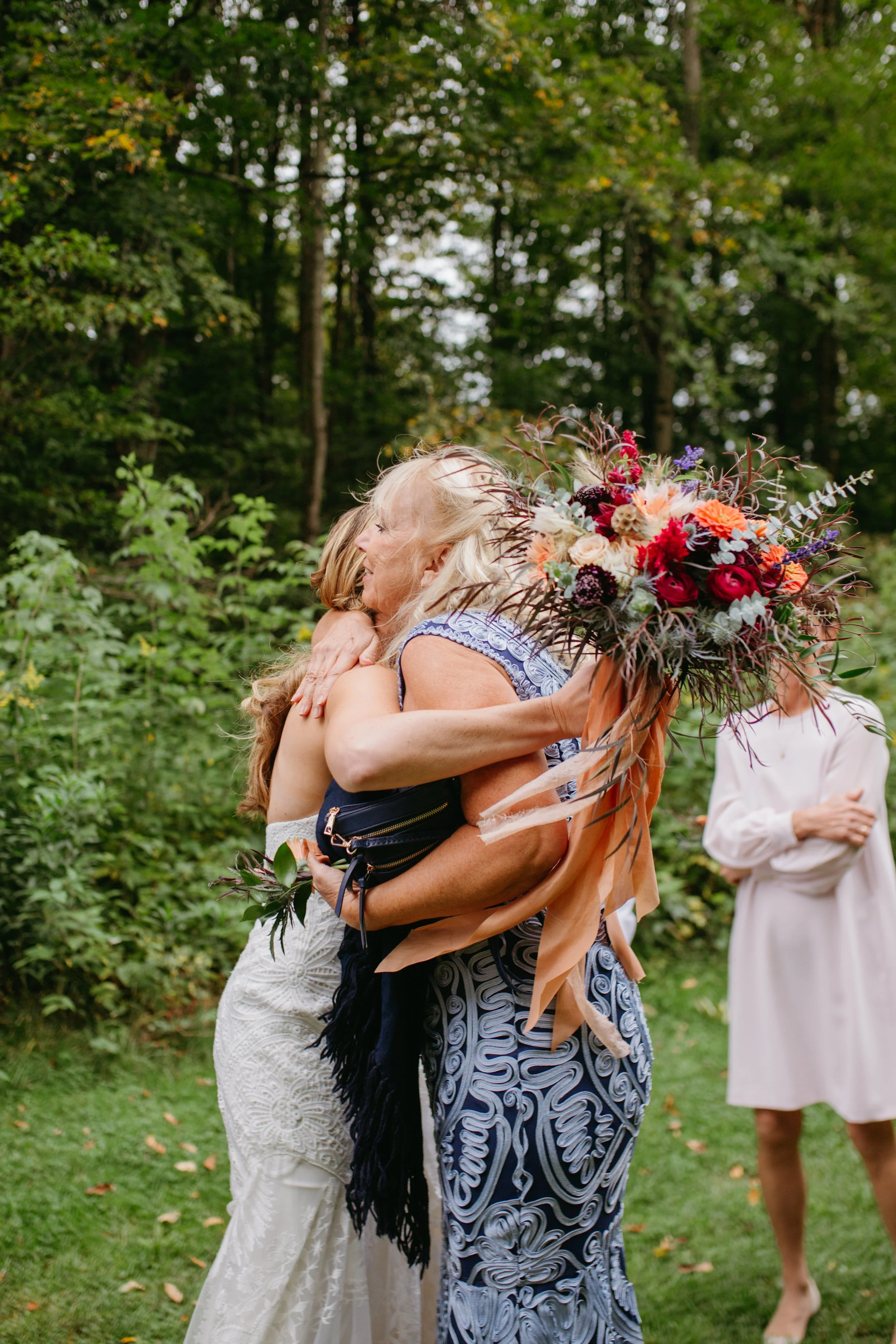 Two women hugging outdoors in a green wooded area, one holding a large bouquet of colorful flowers, while another woman dressed in white observes in the background.