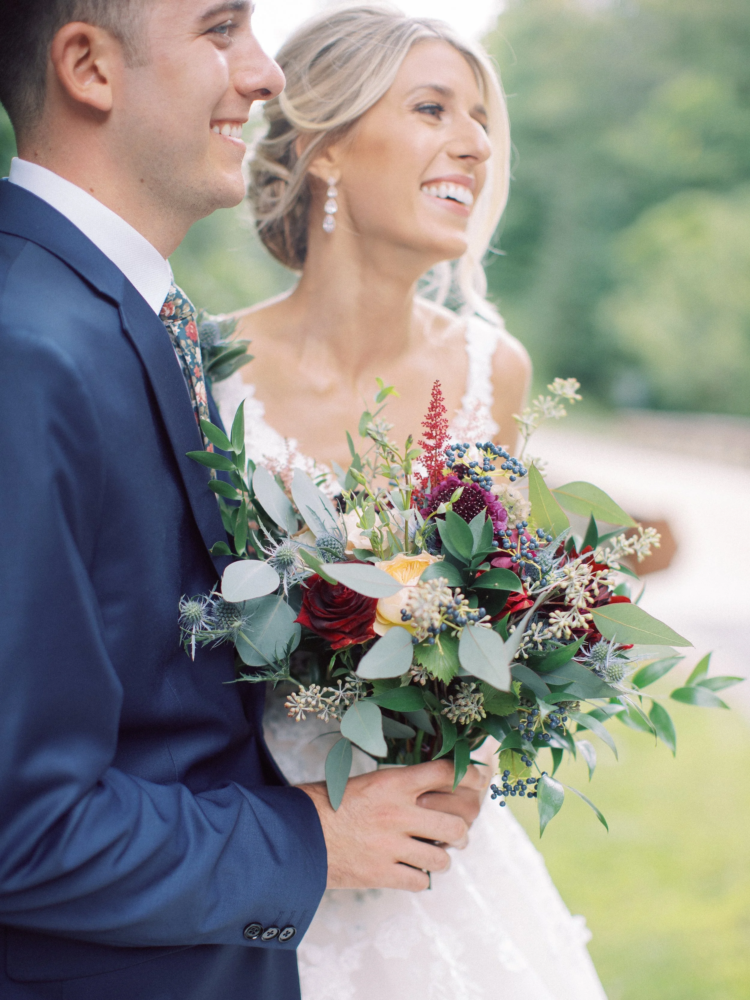 A bride and groom smiling at each other, with the groom in a navy suit and the bride holding a colorful bouquet, outdoors with a blurred green background.