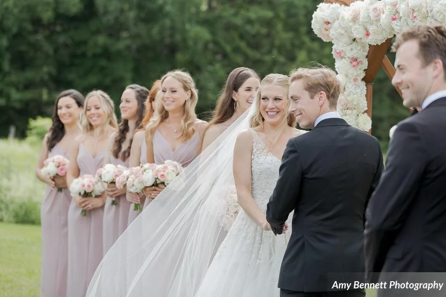 Bride and groom at their outdoor wedding ceremony, with bridesmaids holding bouquets standing behind them, and a decorated flower arch in the background.