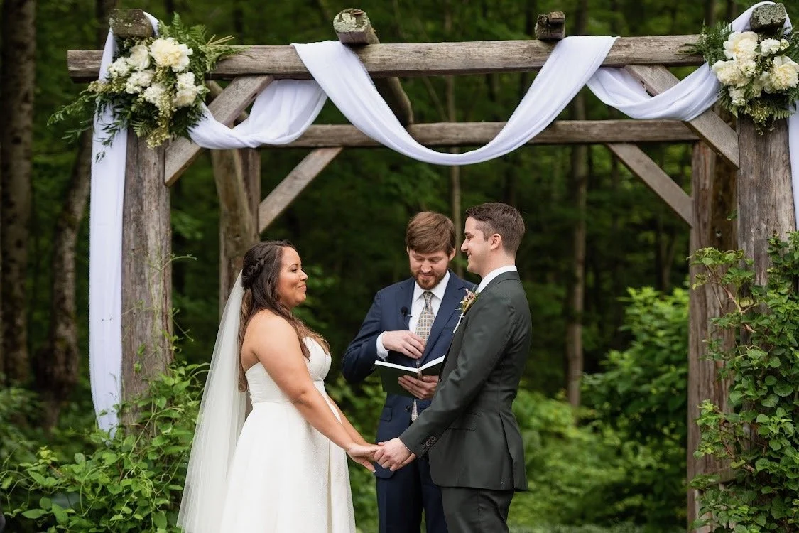 A couple getting married outdoors, holding hands, standing under a wooden arch decorated with white flowers and fabric, with a minister officiating.