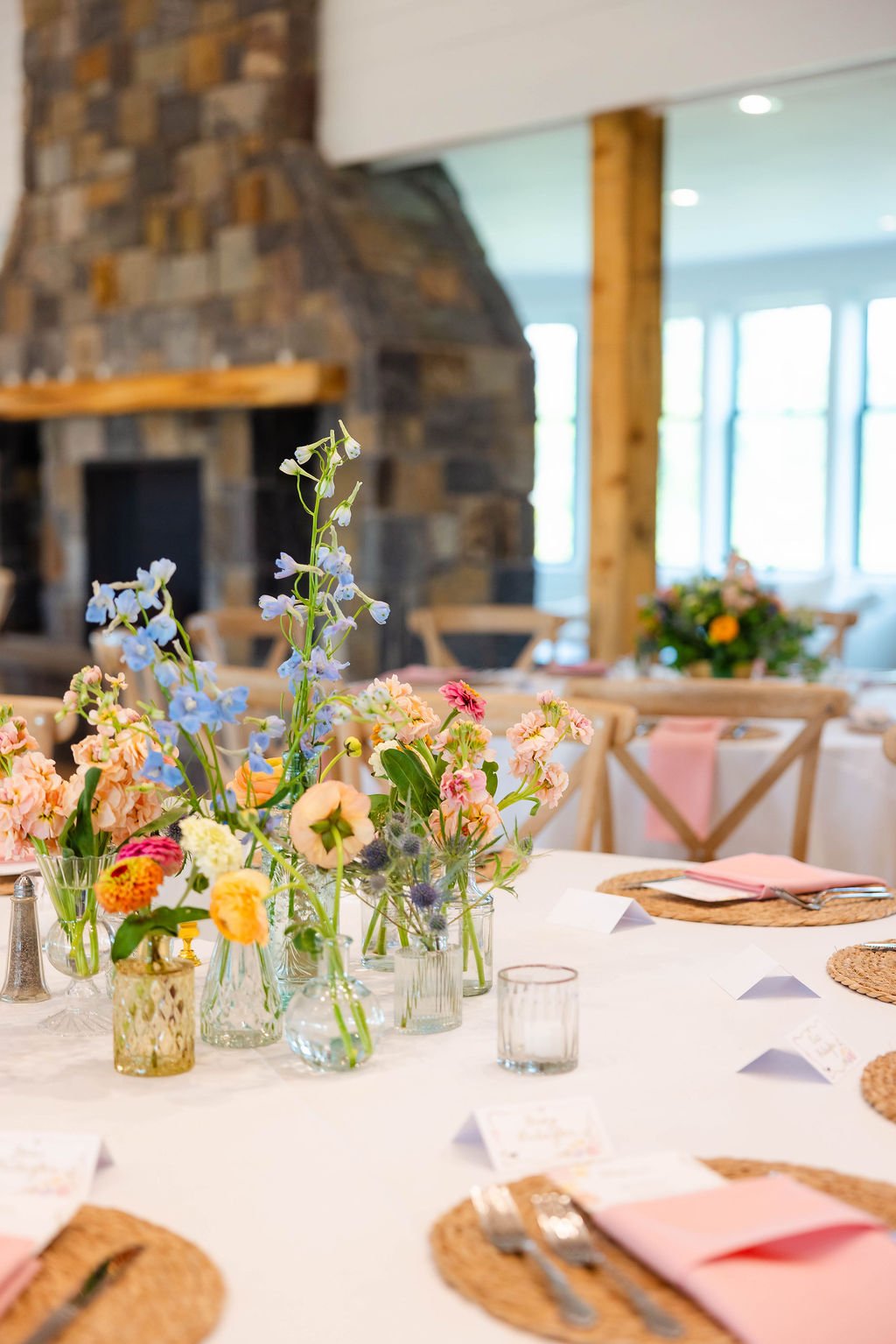 A table decorated with colorful flowers in glass vases, pink napkins, and tableware, set in a rustic indoor setting with a stone fireplace and large windows in the background.