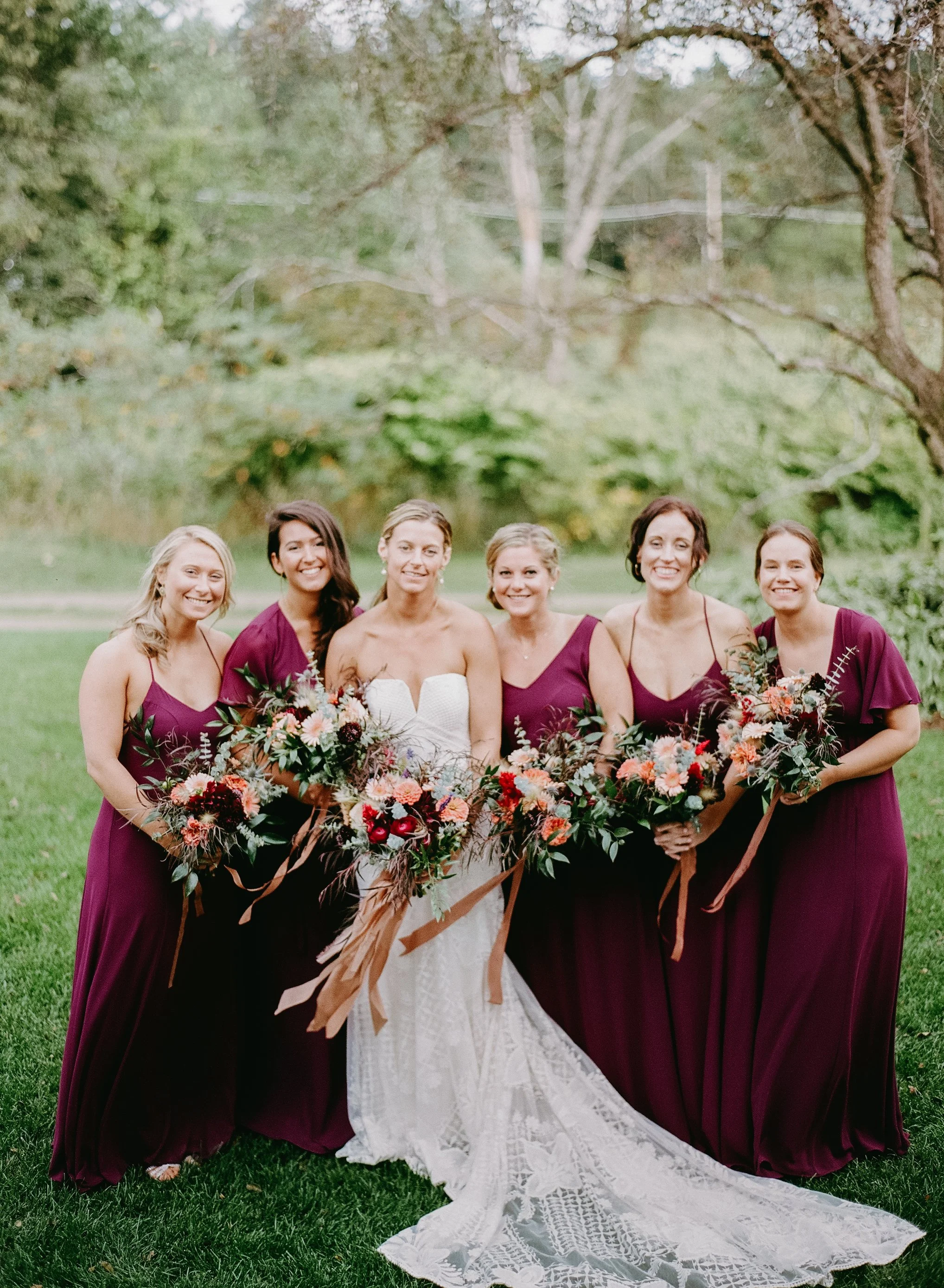 A bride in a white lace dress and six bridesmaids in matching burgundy gowns holding bouquets, standing outdoors on green grass with trees in the background.