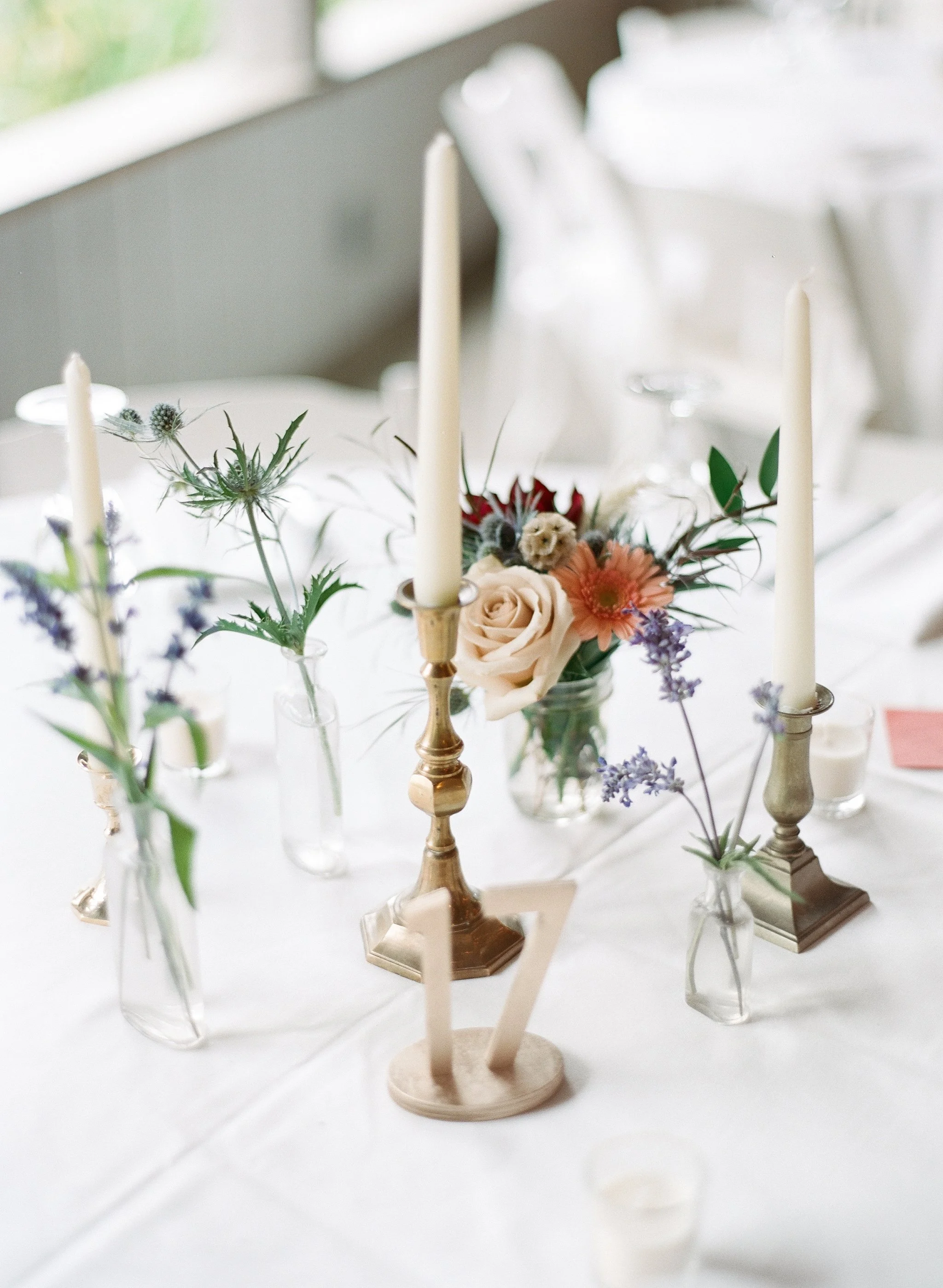 A table decorated with various flower arrangements in small glass vases, with tall white candles in gold candle holders, set on a white tablecloth in a bright room.