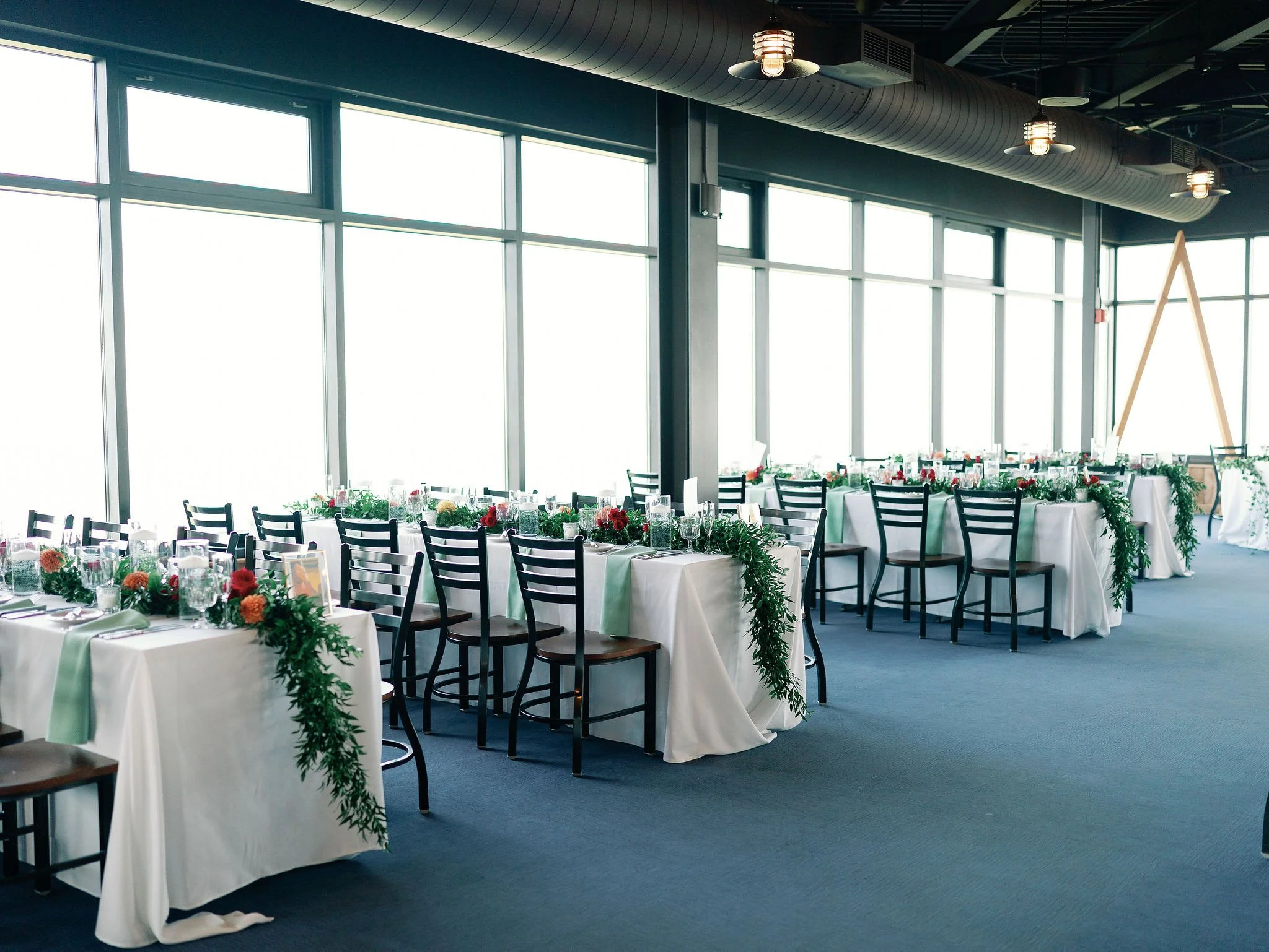 Elegant banquet hall with large windows, decorated tables with white tablecloths, floral centerpieces, wine glasses, and black chairs. Modern lighting fixtures hang from the ceiling.