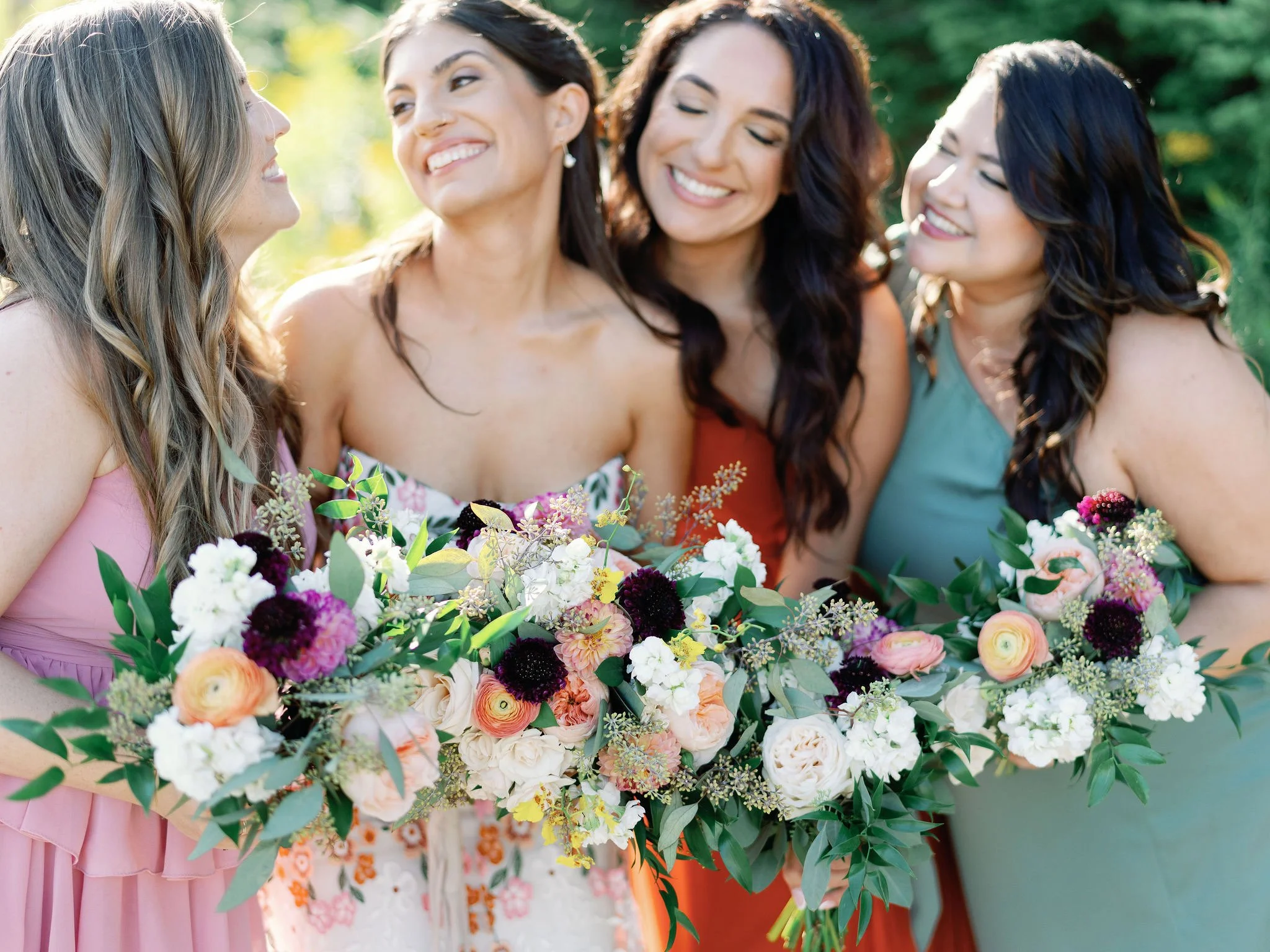 Four women smiling and holding a large bouquet of flowers outdoors during the day.