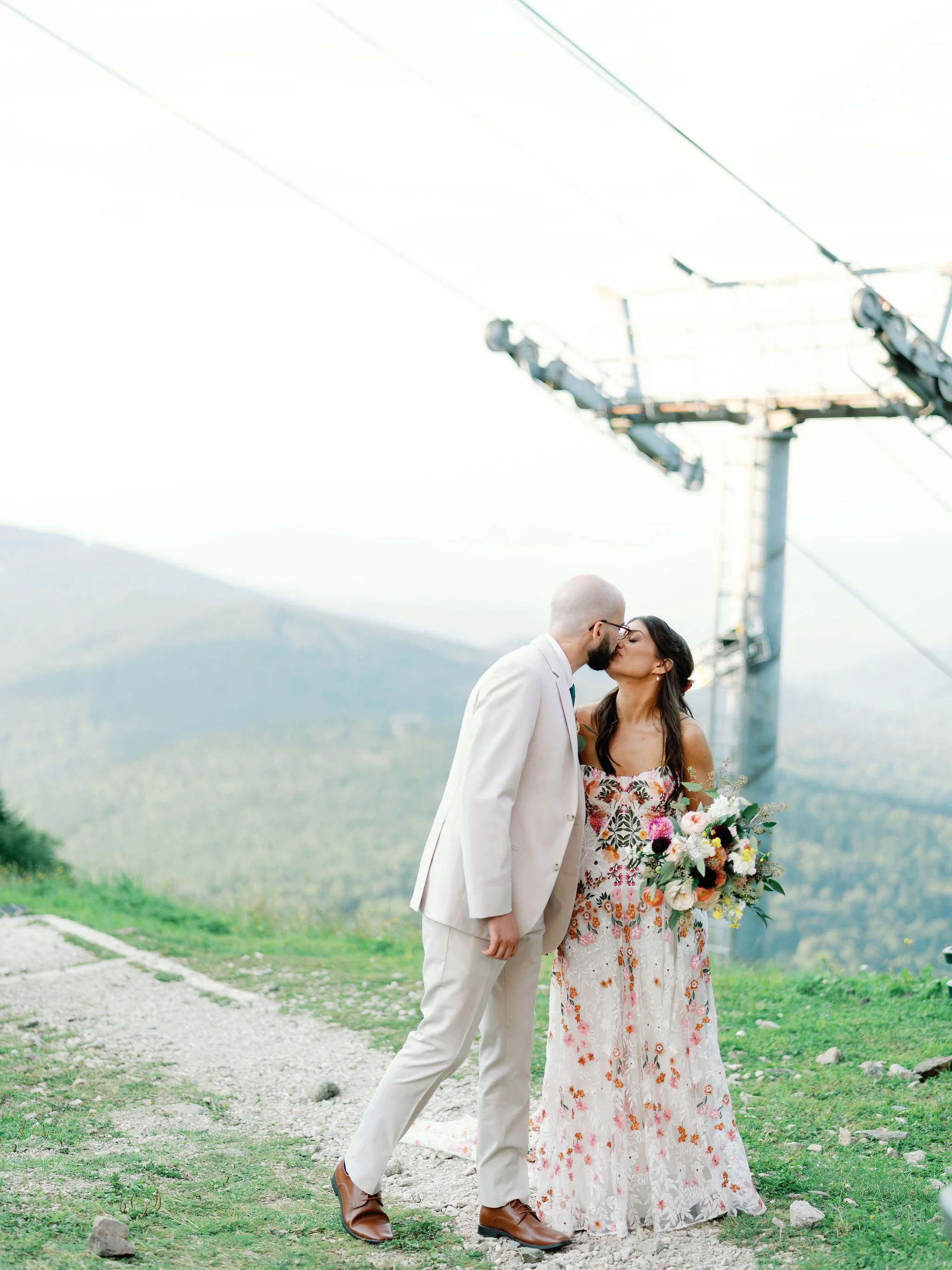 A couple, dressed in wedding attire, sharing a kiss outdoors near a cable car lift with mountains and trees in the background.
