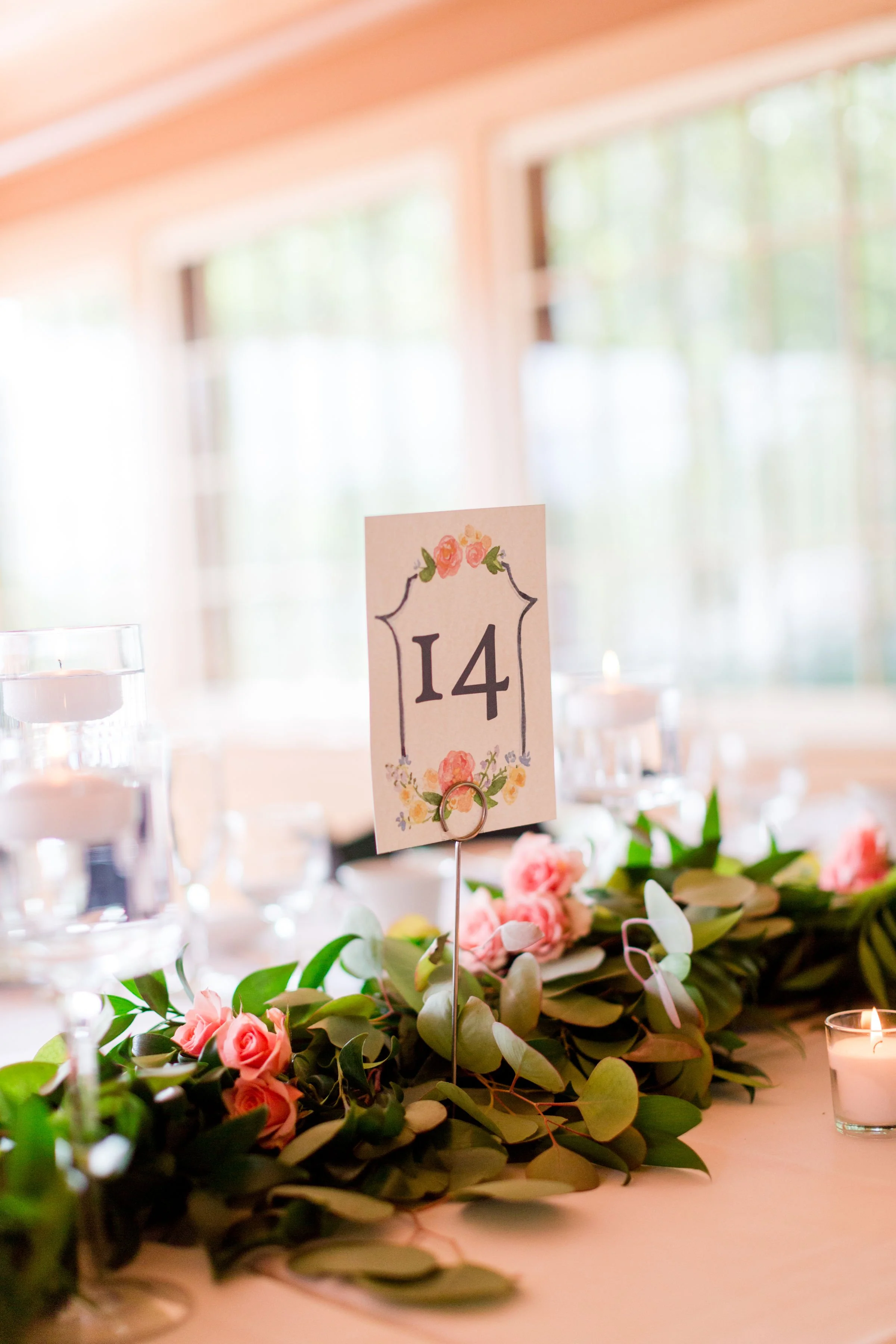 Table centerpiece with pink roses, greenery, and a table number card "14" in a floral design at a decorated event venue.