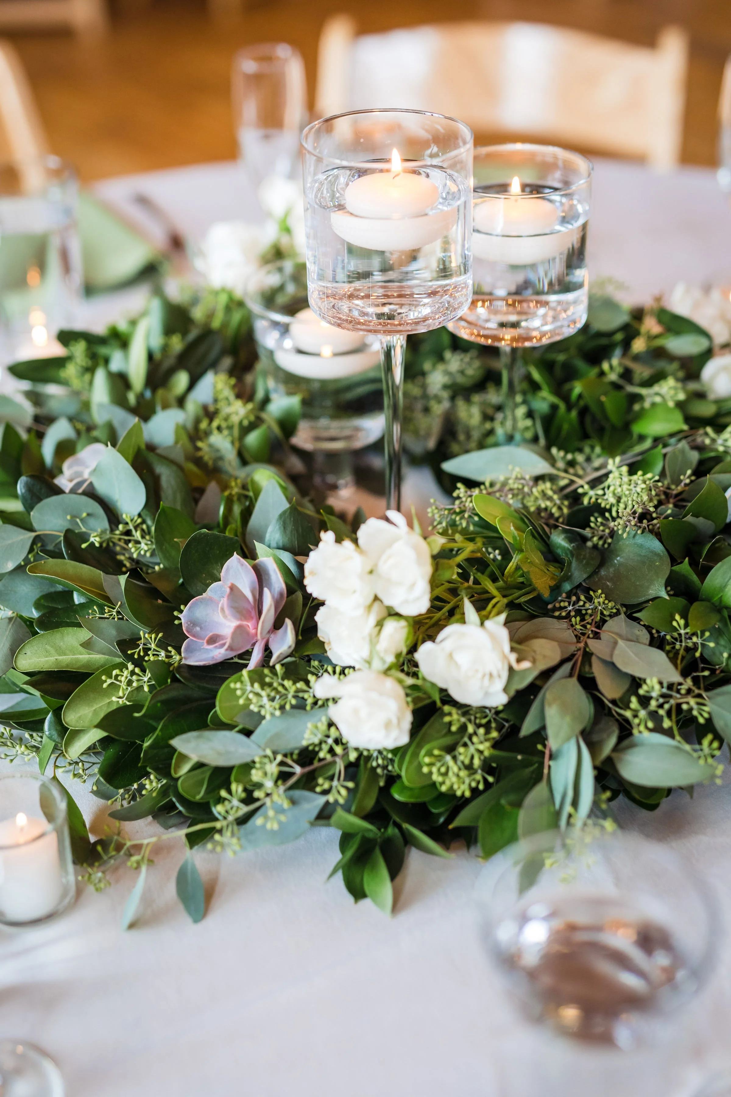 A table centerpiece with floating candles in glass holders surrounded by green leaves and white flowers, on a white tablecloth.