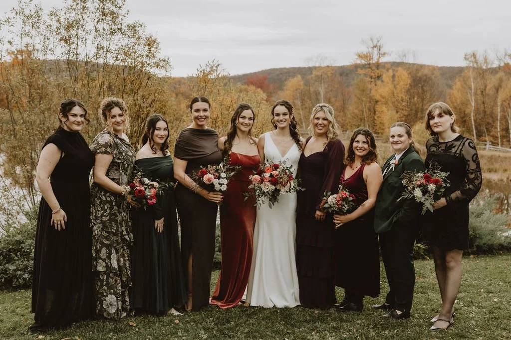 Group of women in formal dresses holding bouquets standing outdoors near a lake with autumn-colored trees in the background.