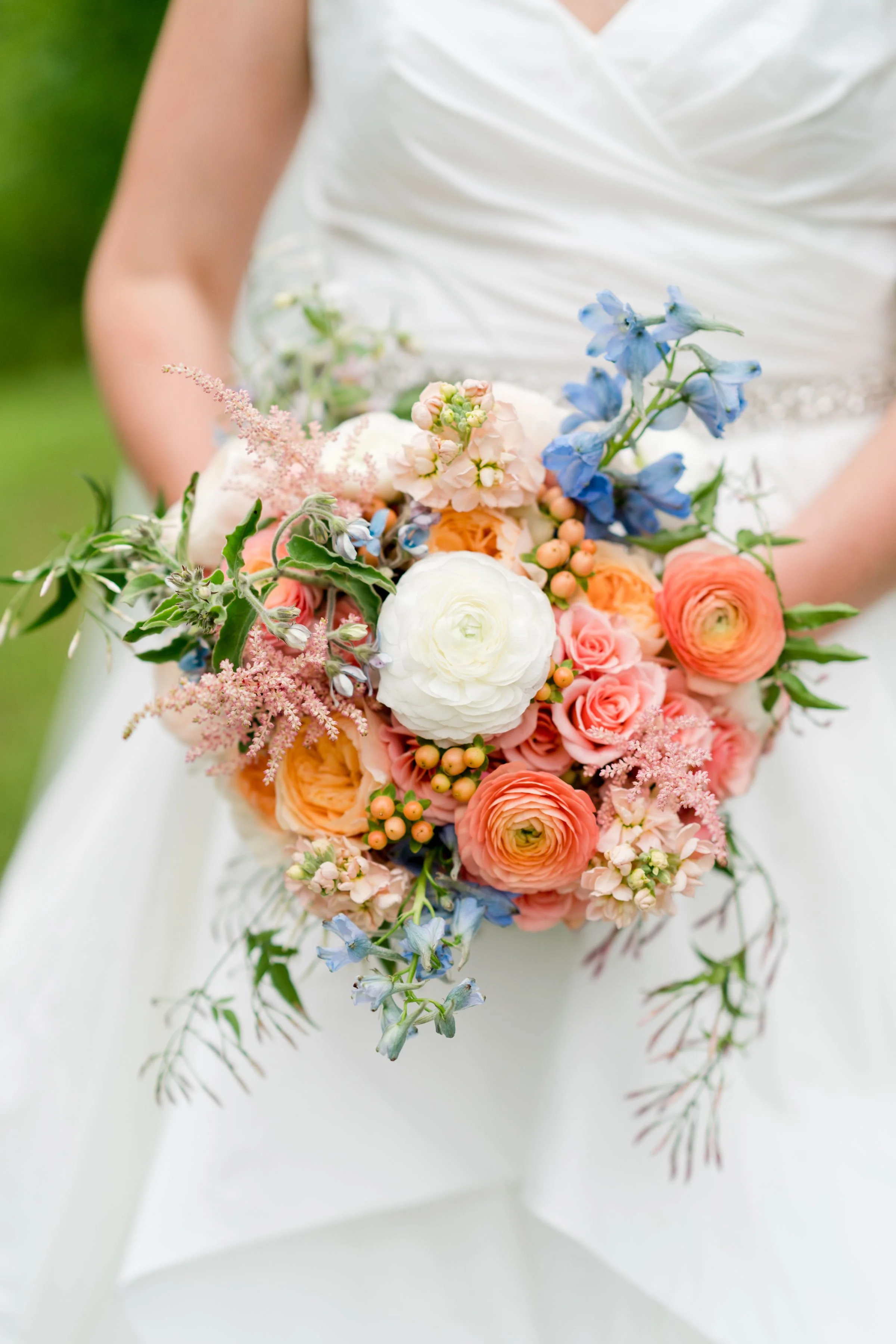 A bride holding a colorful bouquet of flowers including pink roses, white ranunculus, blue delphiniums, peach spray roses, and greenery, standing on a blurred green outdoor background.