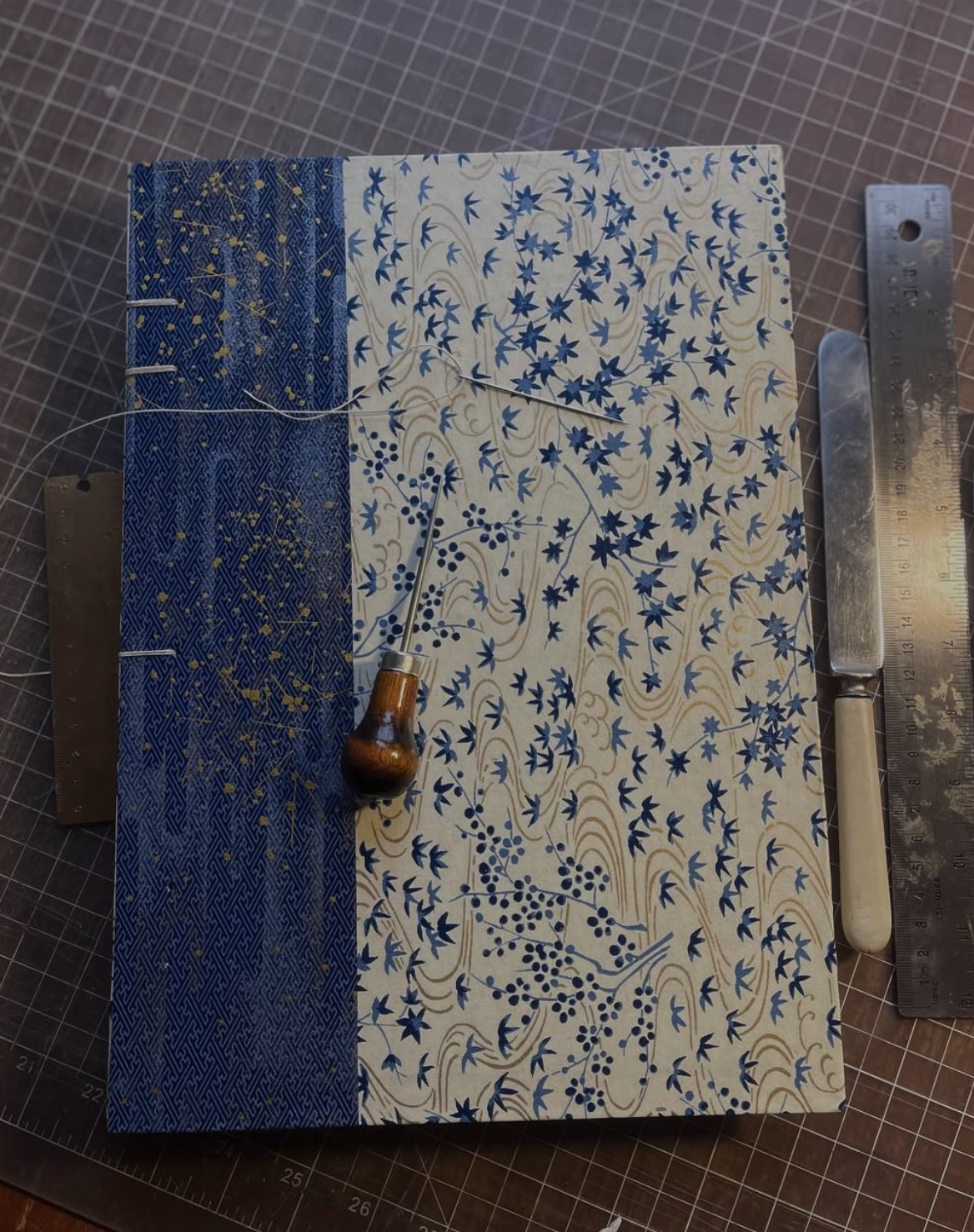 A crafting workspace showing a decorated paper, a pin, a ruler, and a craft knife on a cutting mat.