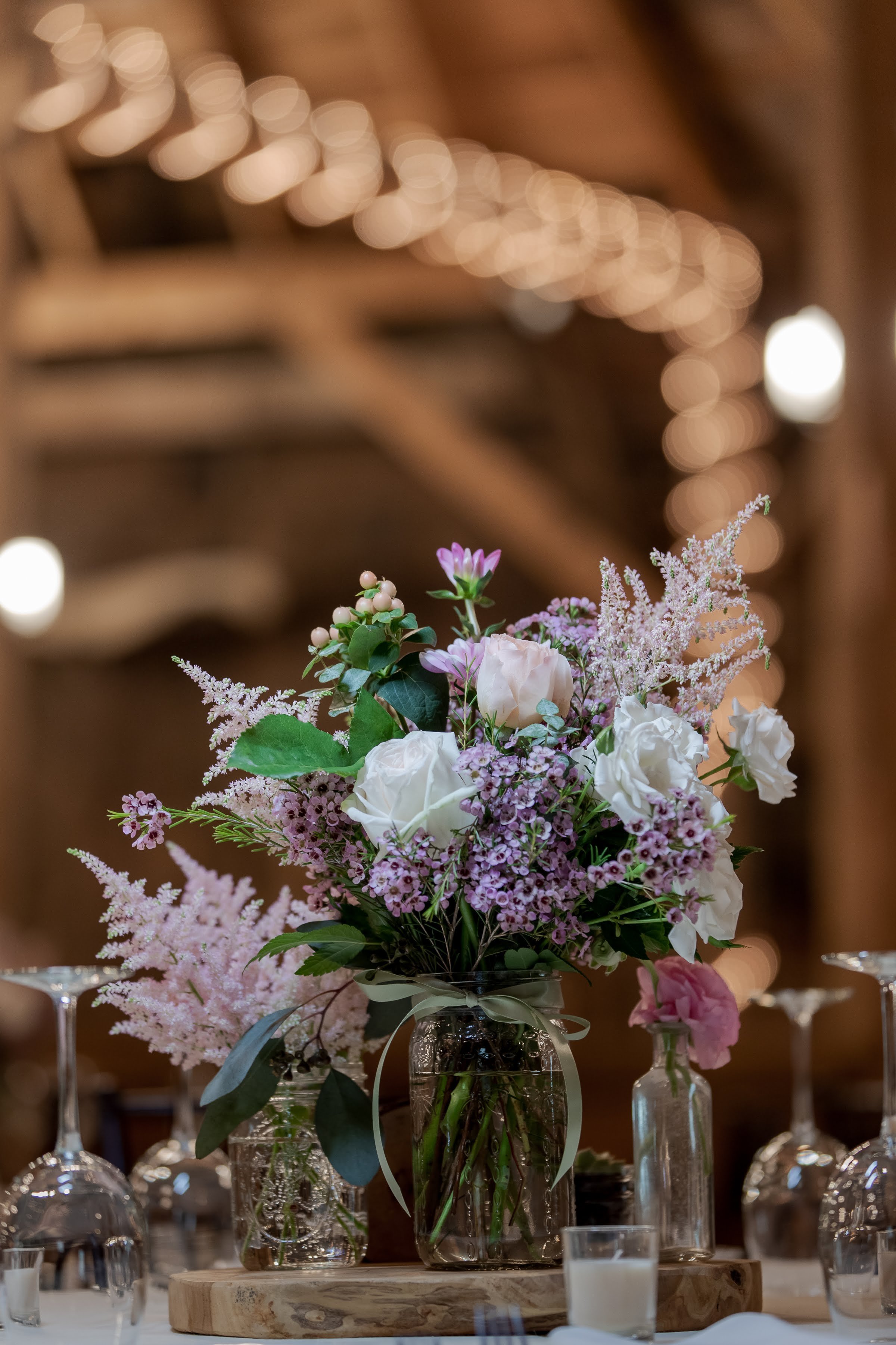 A large floral centerpiece with white roses, pink and purple flowers, and greenery in a glass vase on a wooden table, with soft warm lighting and bokeh in the background.