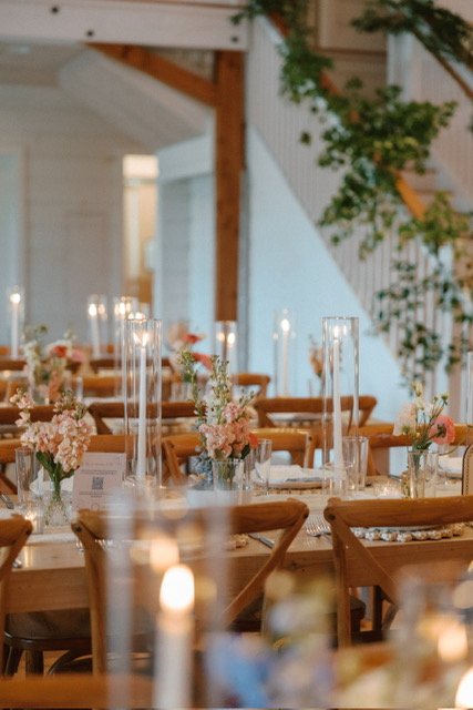 Elegant dining setup with candlelit centerpieces, pink flowers in vases, and wooden chairs in a decorated indoor venue with a staircase and greenery.