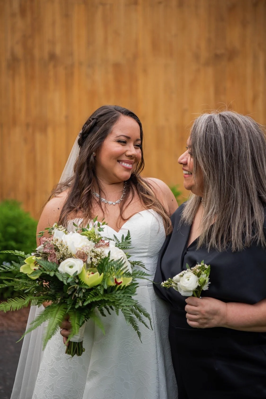 Two women, one in a wedding dress and the other in black, smiling and holding bouquets of flowers, standing close together.