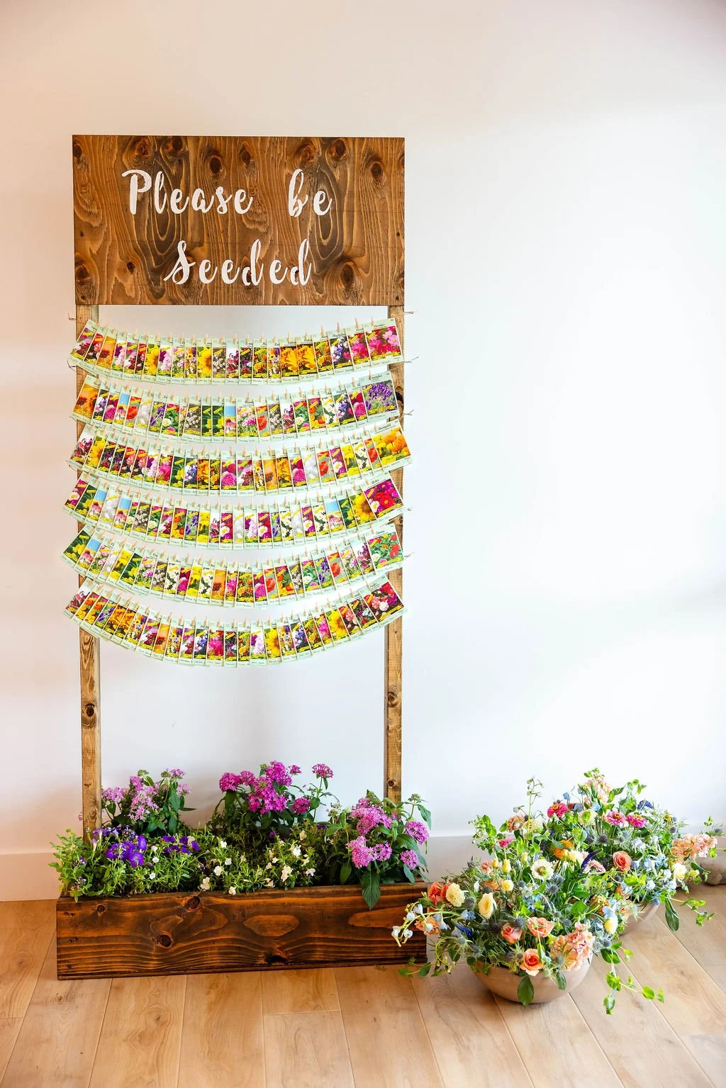 Display of colorful flower seed packets on a wooden stand with a sign that reads 'Please be seedled.' The stand is decorated with pink, purple, and white flowering plants in a wooden planter box and a large white bowl.