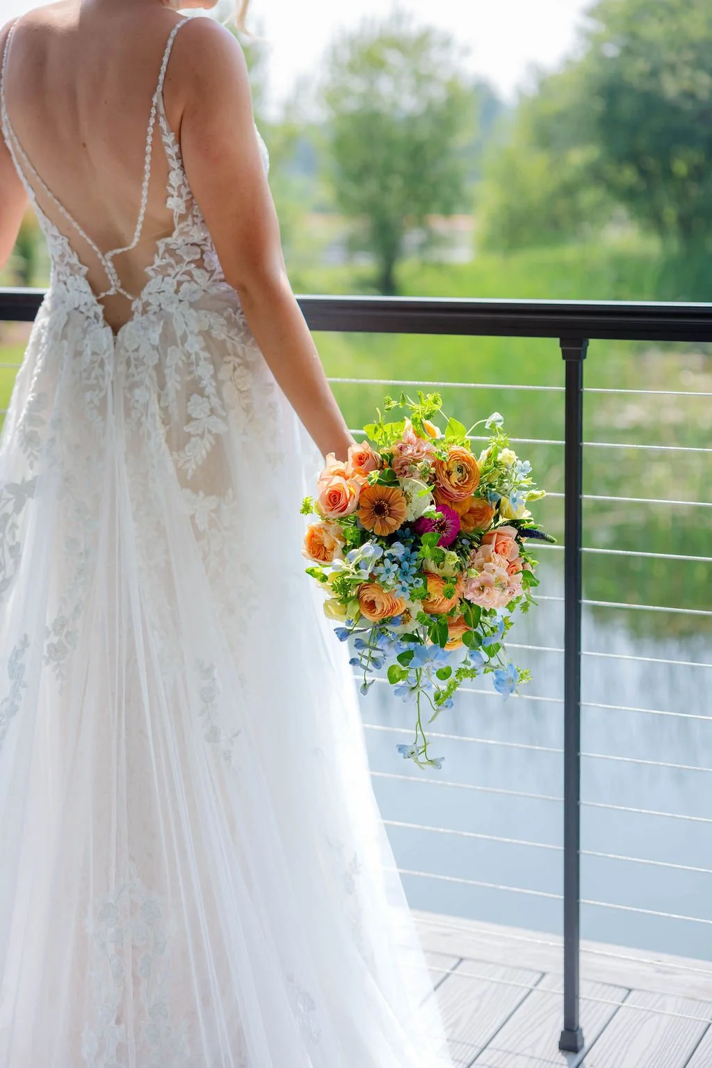 A bride in a white lace wedding dress holding a colorful bouquet of flowers, standing by a balcony overlooking a green outdoor landscape.