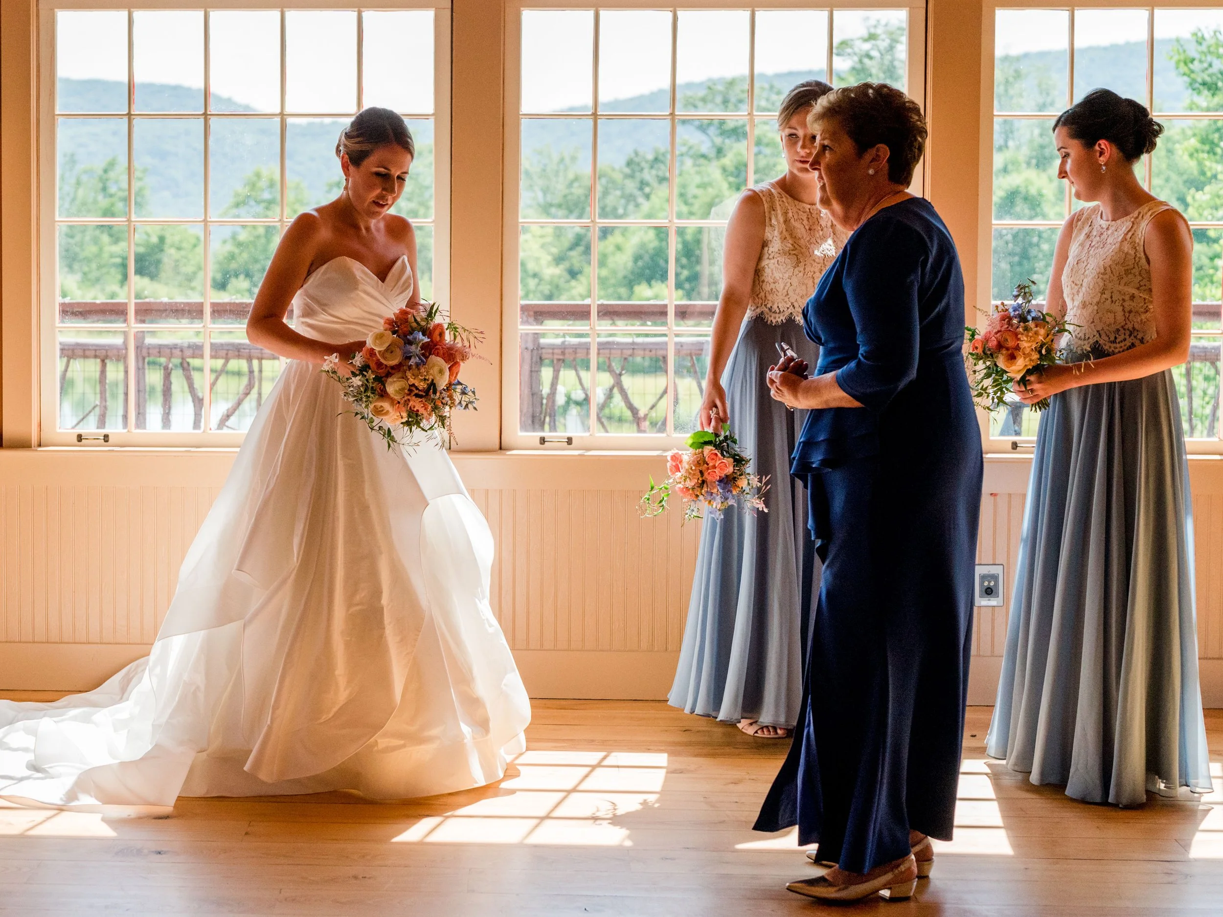 A bride holding a bouquet inside a room with large windows overlooking a green landscape, with three women and a woman in a blue blazer standing nearby.